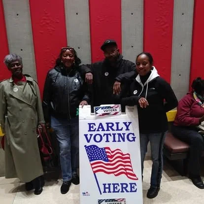 Four individuals gathered around an early voting sign with an American flag, indoors with a wooden wall background.