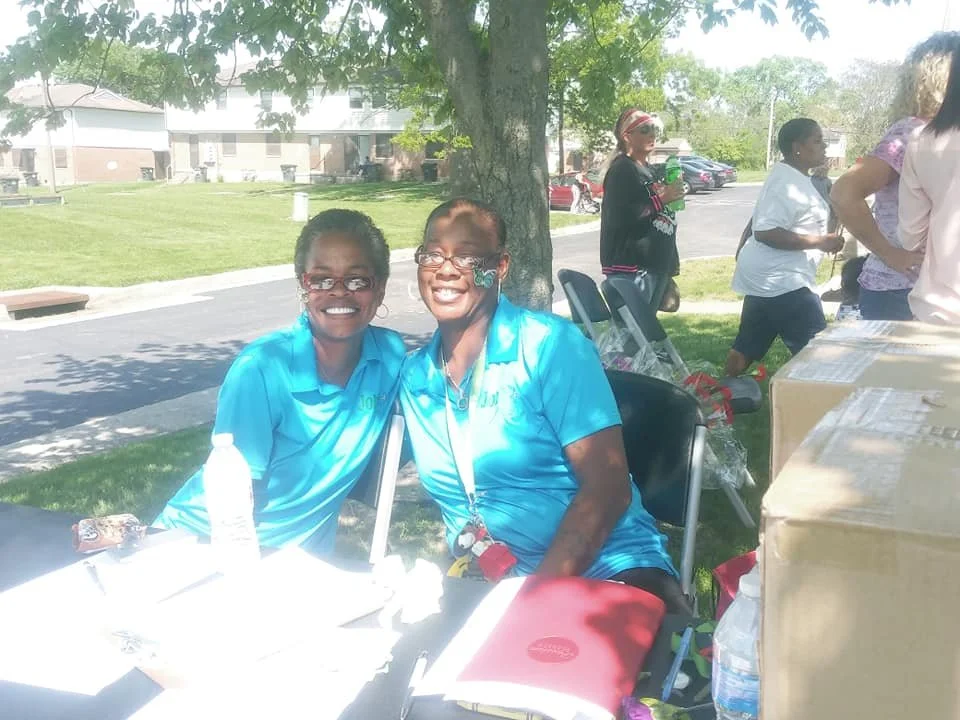 Two women in blue shirts sitting under a tree at an outdoor event, smiling into the camera. Five other people are visible in the background, some standing near tables and others holding bottles.