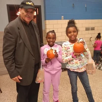 Two young girls smiling and holding pumpkins, with an older man in a suit and cap standing beside them at an indoor event.