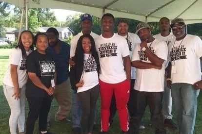 Group of nine diverse people standing together outdoors under a tent, smiling, with some wearing matching T-shirts.