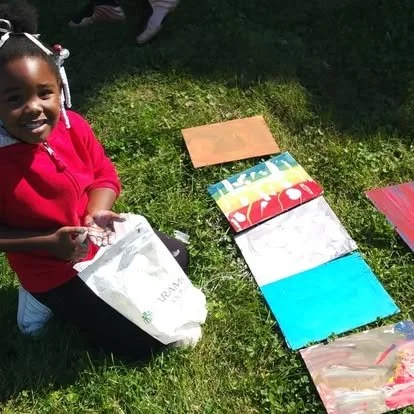 A young girl in a red hoodie kneels on grass, smiling beside her colorful painted canvas sheets laid out on the grass.