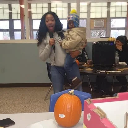 Woman holding a baby and speaking into a microphone at an indoor event, with pumpkin decoration on table in foreground.