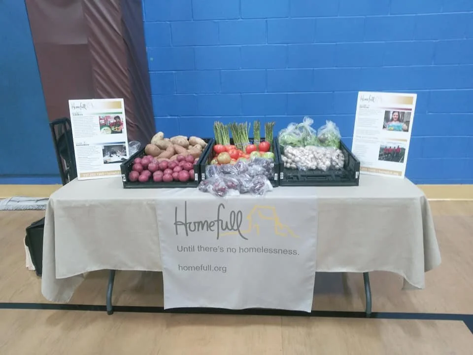 Table with fresh vegetables including potatoes, radishes, green onions, tomatoes, garlic, and garlic bulbs, with informational signs and a banner for Homefull organization promoting homelessness prevention.