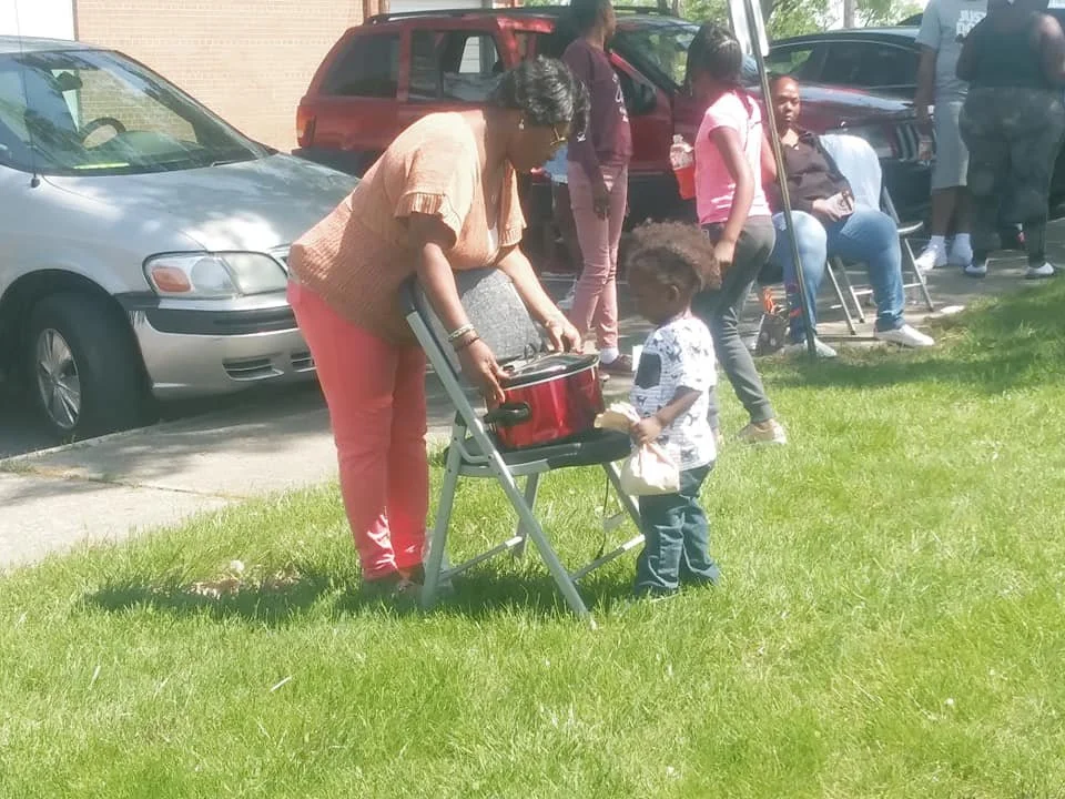 A woman and a small child are outdoors near a parking lot. The woman is leaning over a folding chair with a red cooler on it, while the child looks at the cooler. Several other people are in the background, some sitting and some standing, with cars p