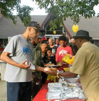 People gathered outdoors at a community event, standing in line to receive food from a man serving at a table covered with aluminum trays and yellow packaged items, with trees and a building in the background.