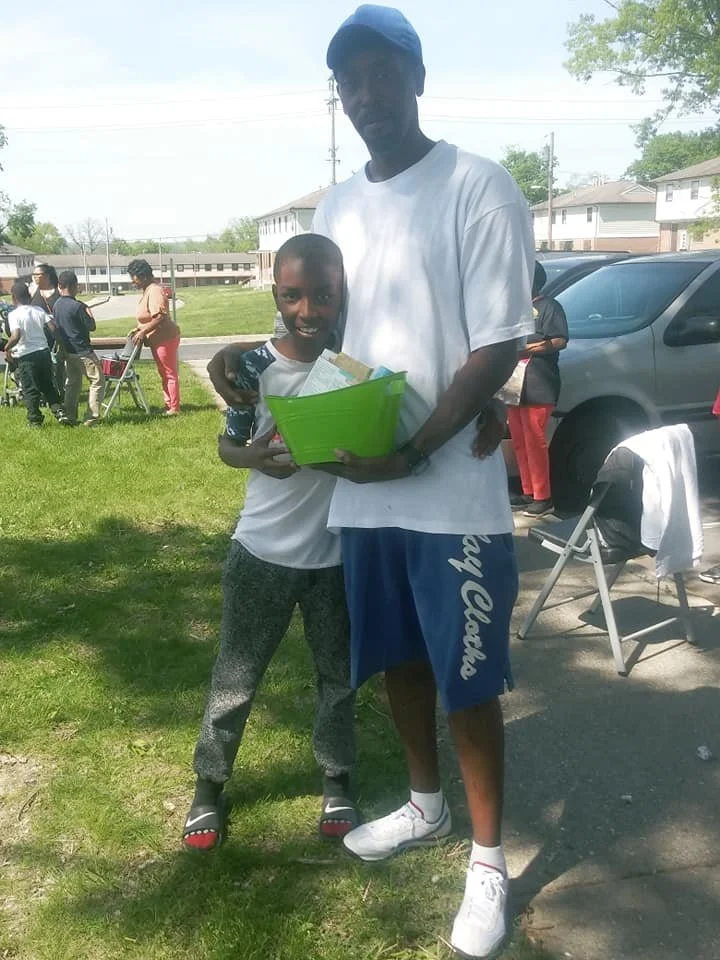 A young boy and a taller man smile and hold a green container filled with papers or gifts at an outdoor event. Several people and cars are visible in the background on a sunny day.