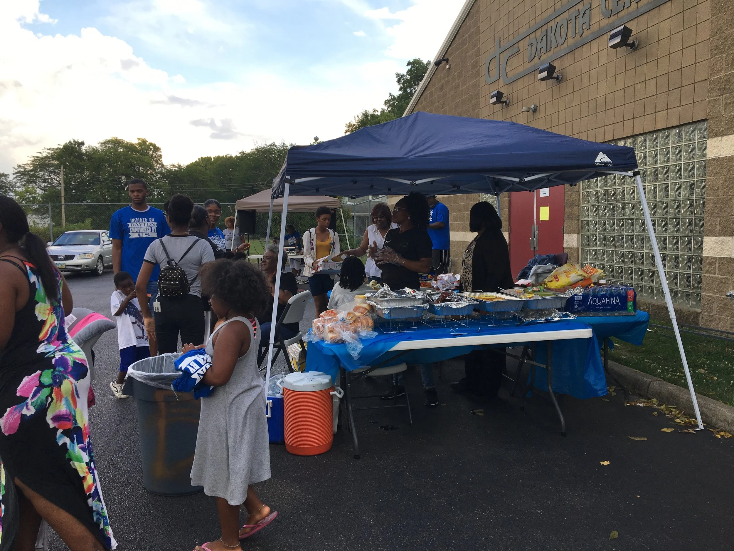 People gathering around a food and snack table set up outside a building with a sign that reads Dakota Center; several people are standing and sitting while a blue canopy is covering the table.