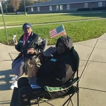 Two people sitting outdoors on foldable chairs, one holding a flag with stars and stripes, and the other holding a drink, with a grassy area and a building in the background.