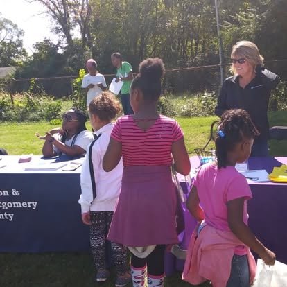 Group of children and adults gathered outdoors at a table during daytime, with trees and grass in the background.
