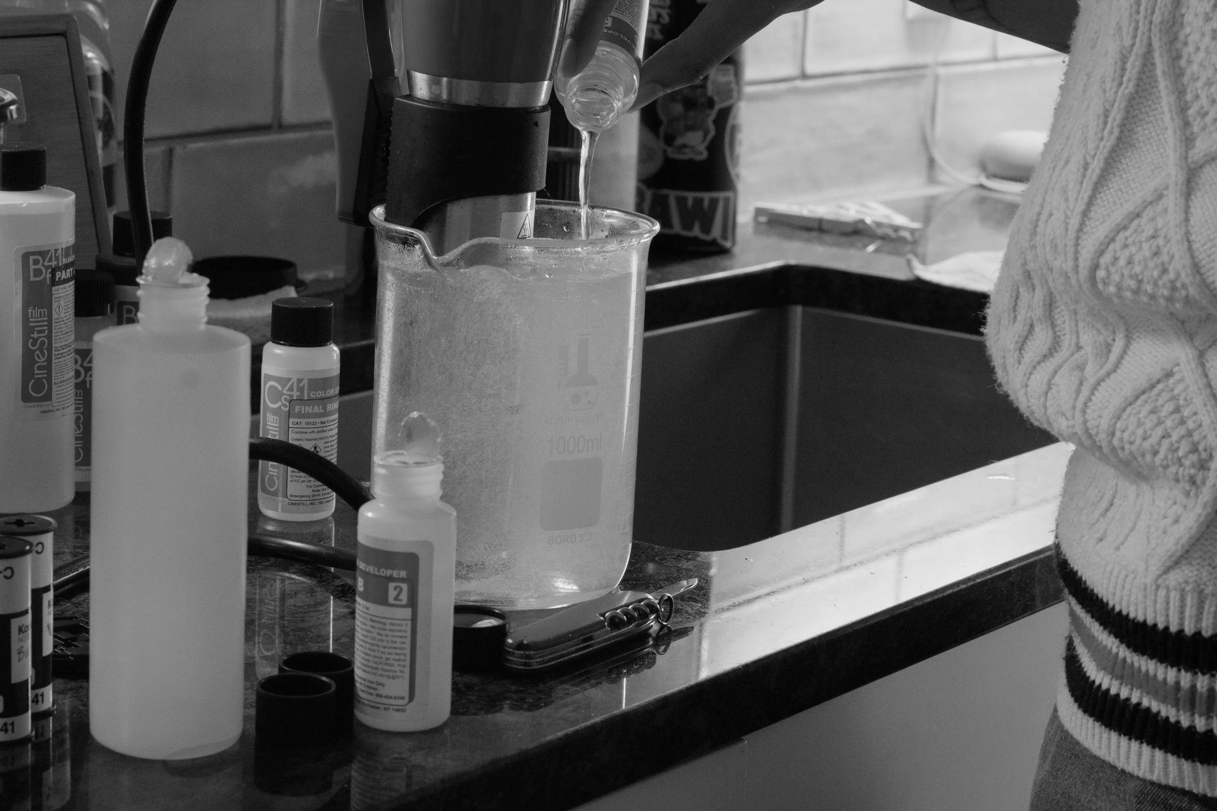A person wearing a striped sweater pouring a liquid into a graduated beaker in an at-home film lab, with various bottles and lab equipment on the countertop.
