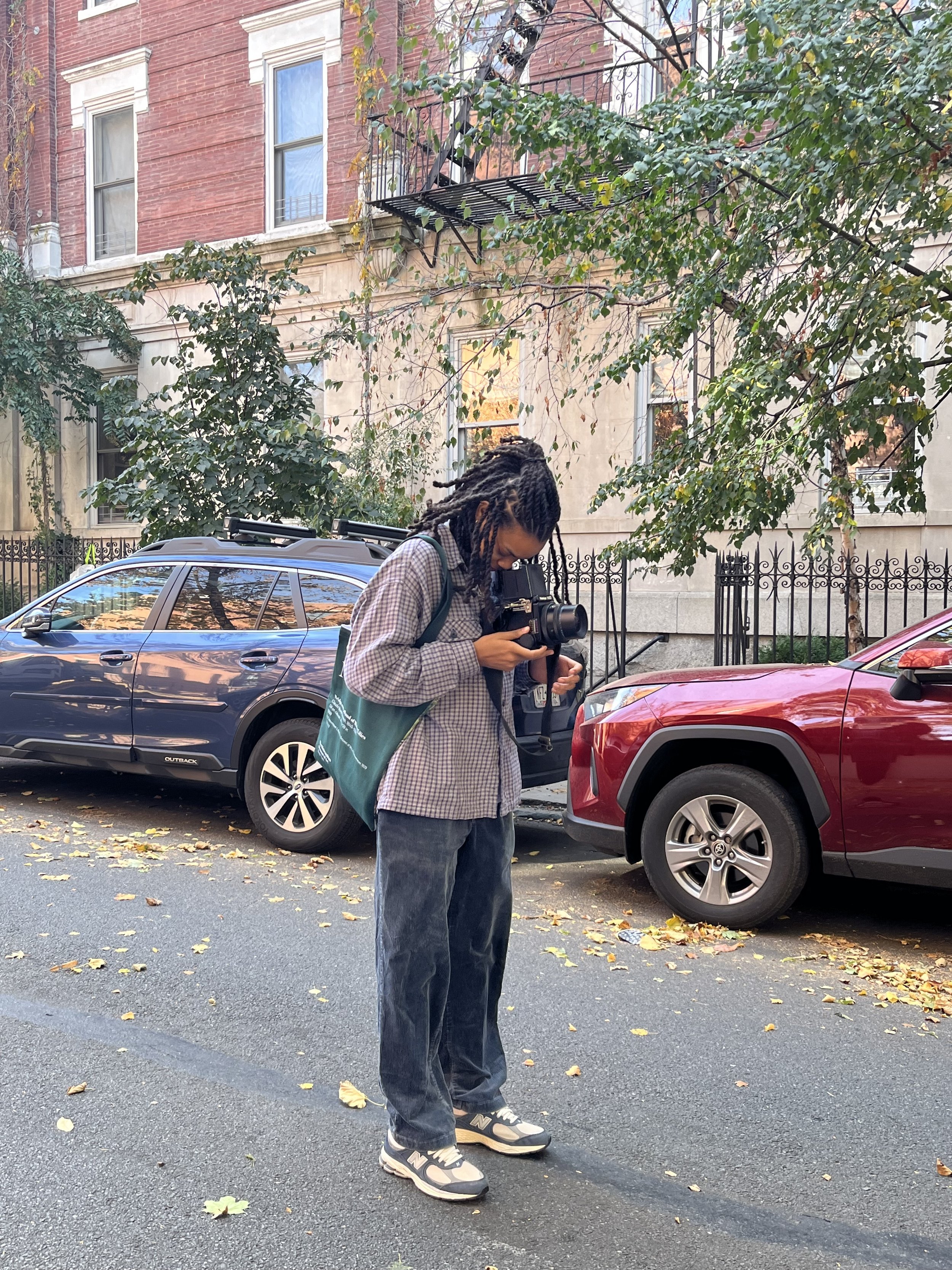 A person with locks wearing a plaid shirt and sneakers is taking a photo, standing on a street with parked cars and fallen leaves, with a building and trees in the background.