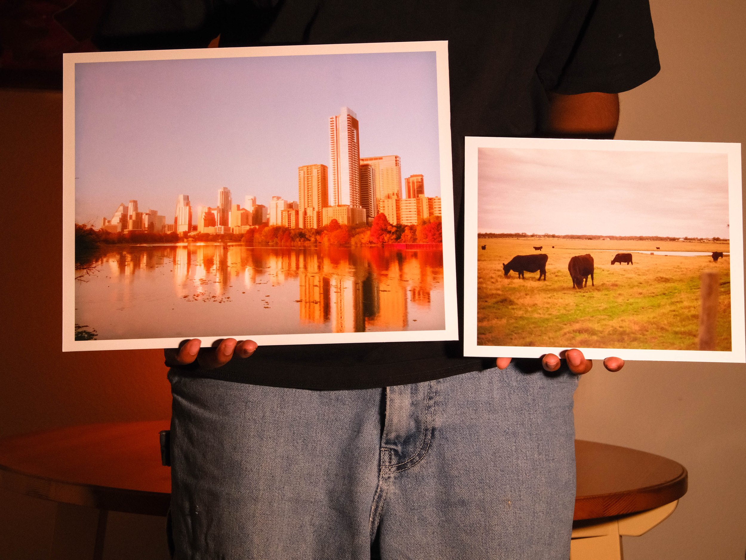 A person is holding two photographs. One photo shows a city skyline with tall buildings and a body of water in the foreground, reflecting the buildings, during sunset. The other photo depicts a rural landscape with cows grazing on a field.
