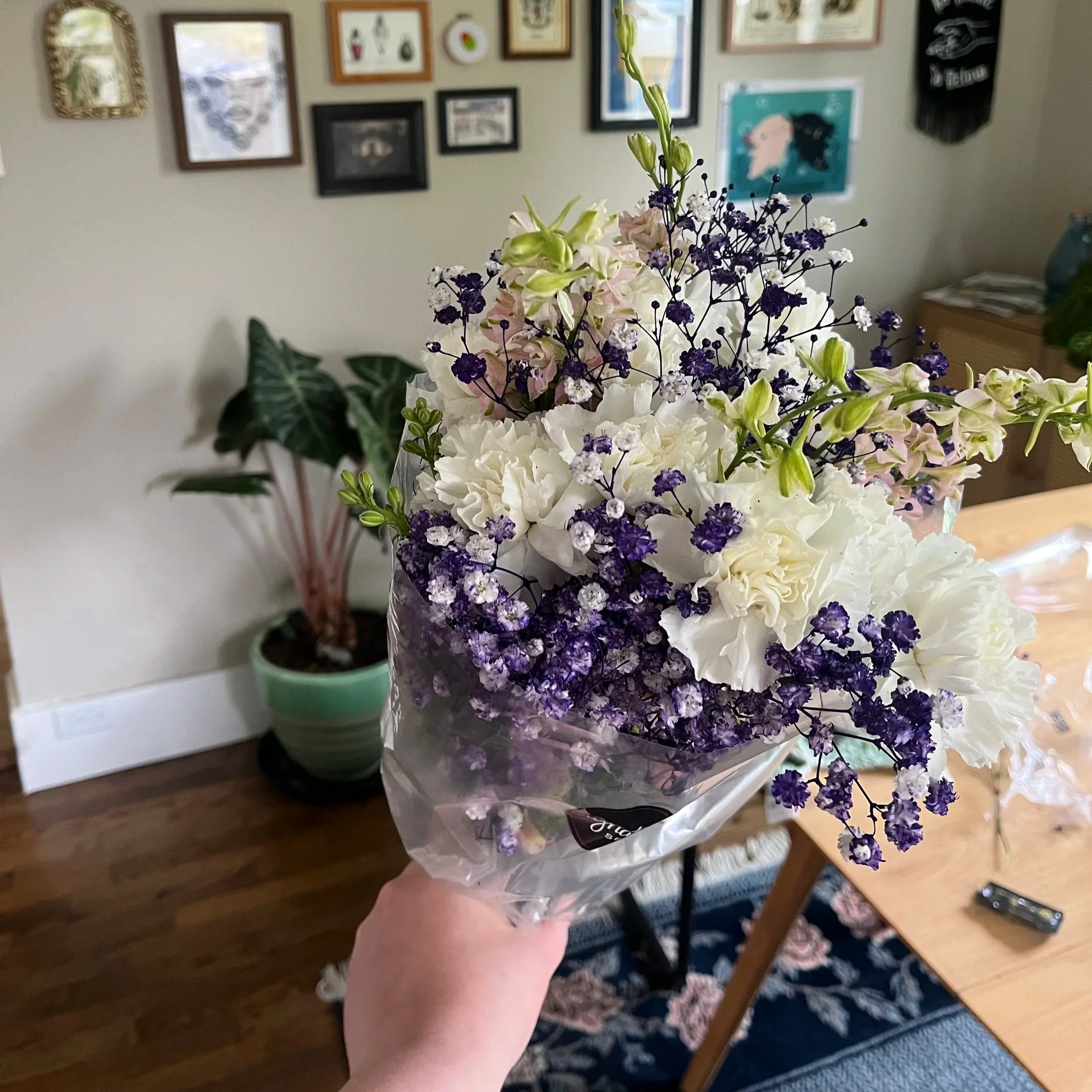 A person's hand holding a bouquet of white and purple flowers, with framed pictures on the wall in the background.