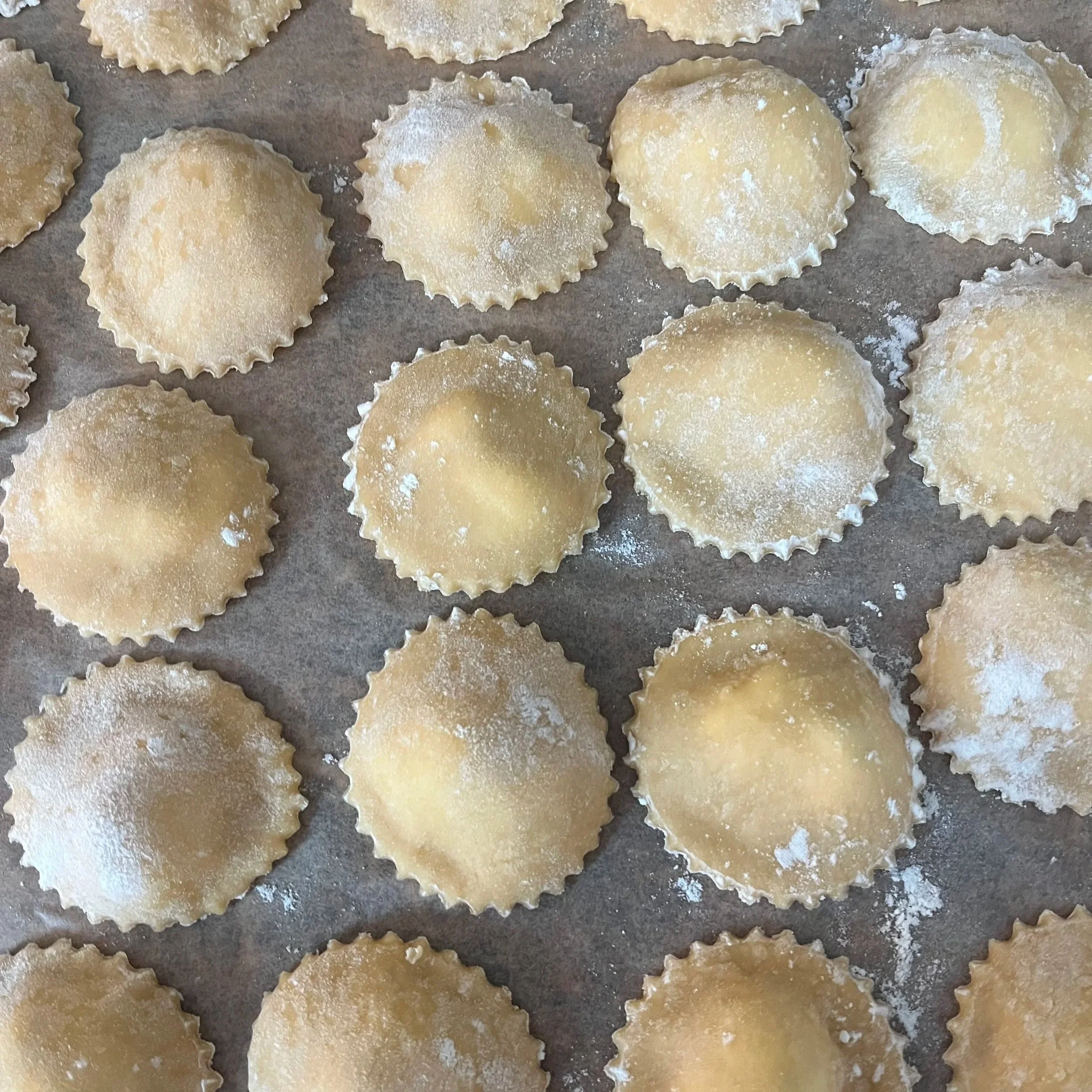 Uncooked ravioli pasta with fluted edges, dusted with flour, on a baking sheet.