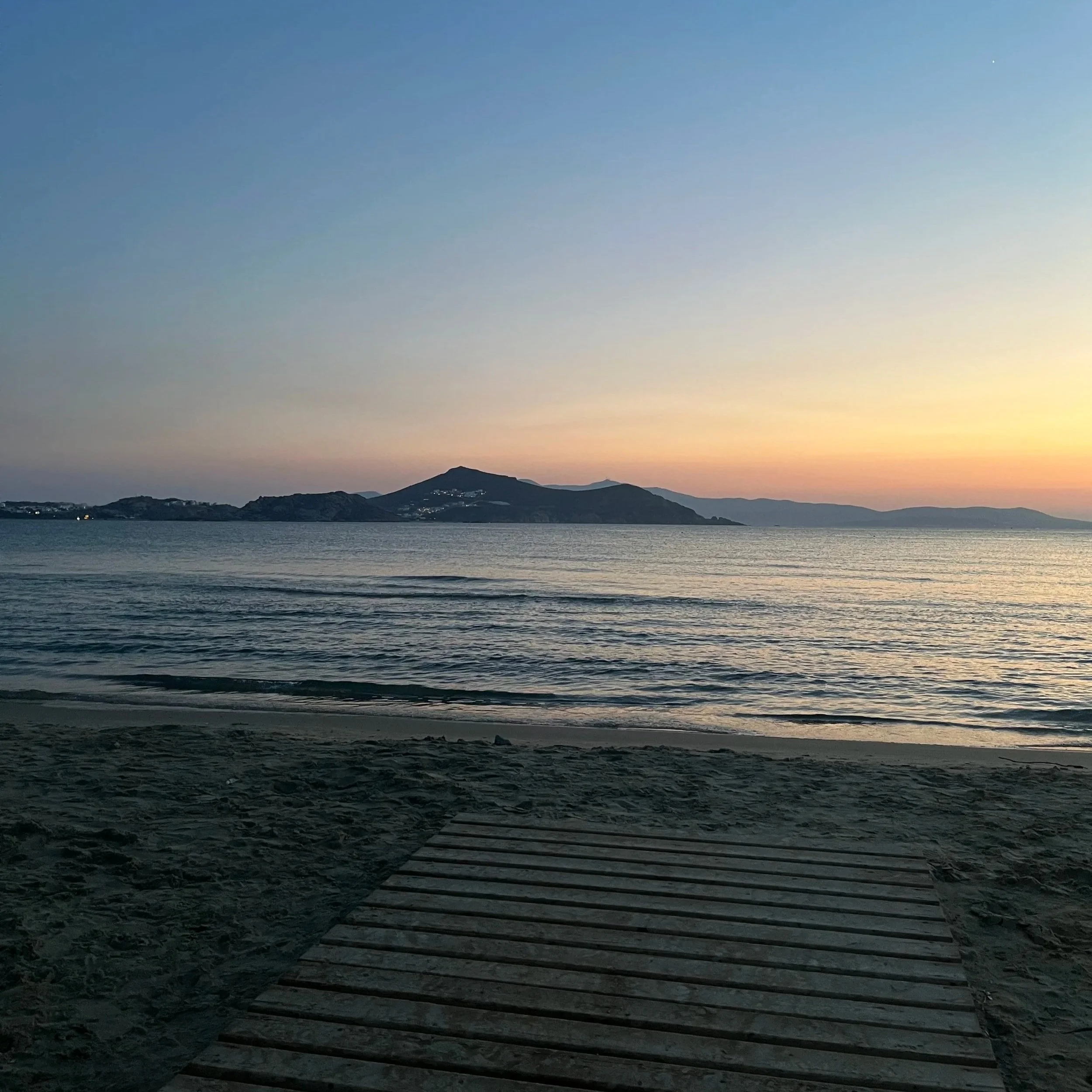 A wooden dock extends into a calm body of water at sunset, with distant mountains and a colorful sky in the background.