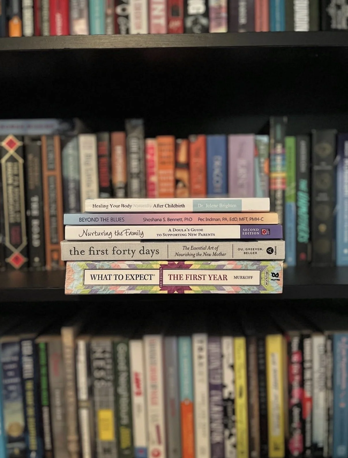 A stack of seven books on a black bookshelf, with colorful book spines visible in the background.