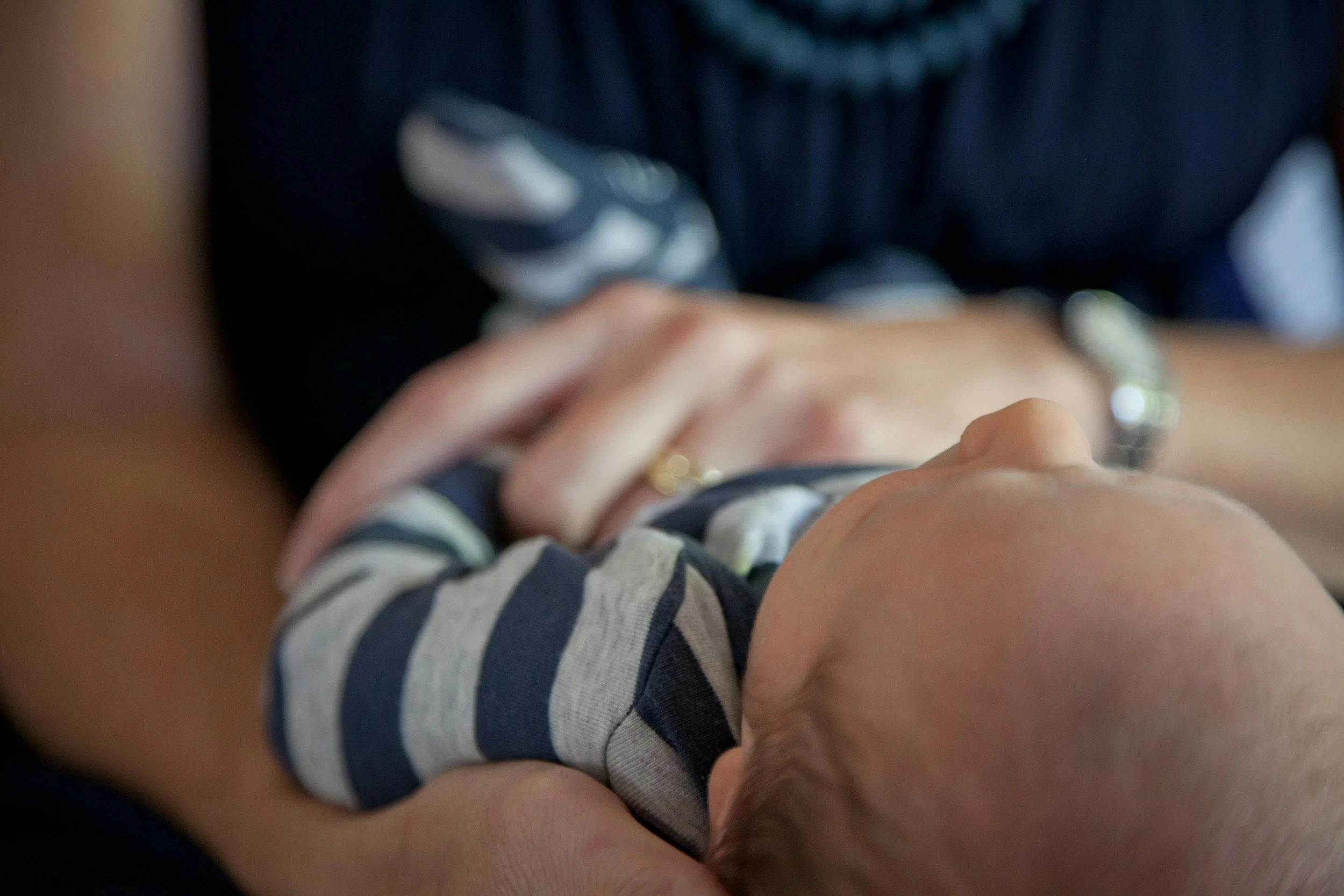 Close-up of a baby lying on an adult's arms, wearing a striped outfit, with the person's hands gently holding the baby.