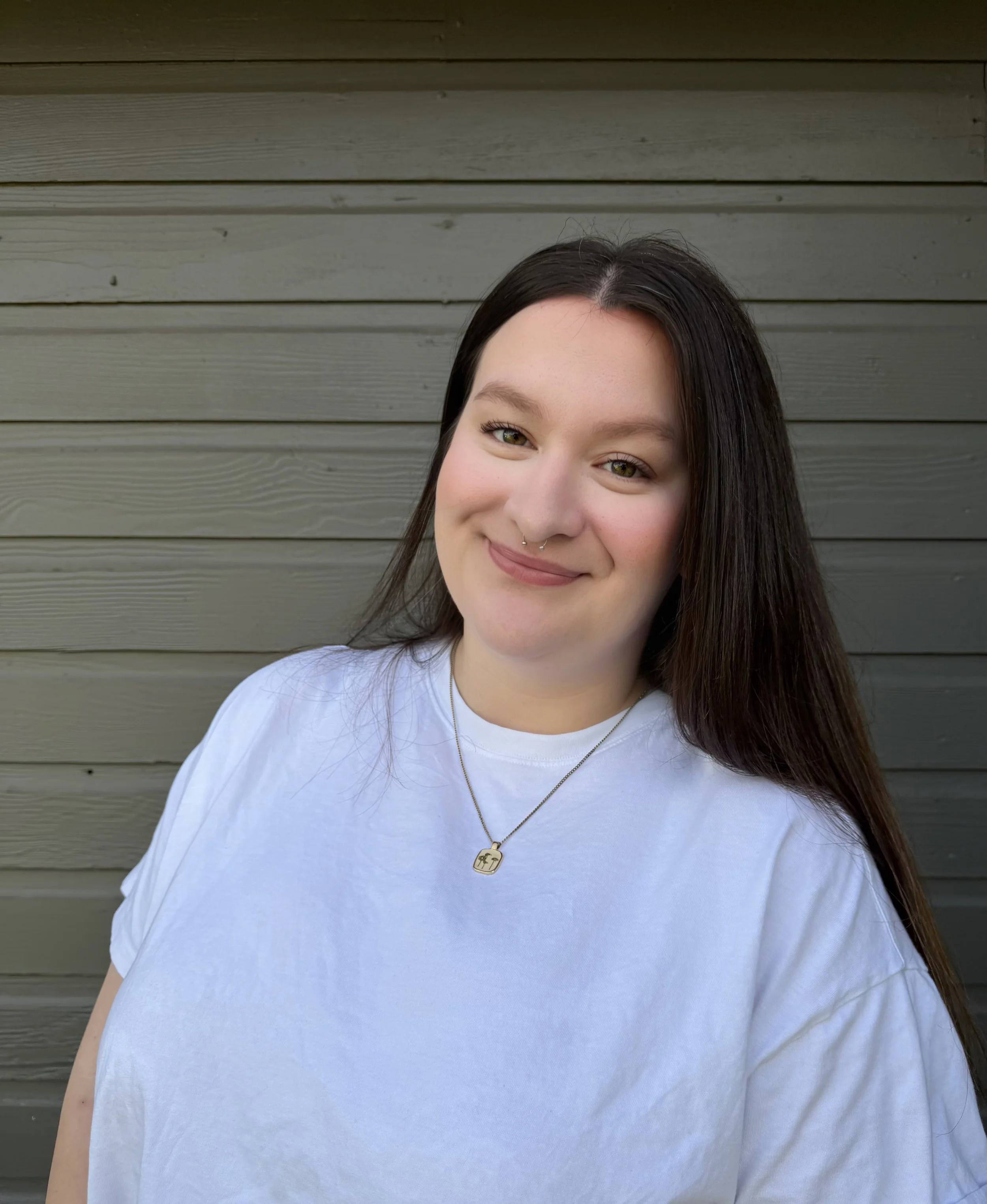 Close-up of a young woman with long dark hair, smiling, wearing a white shirt and a gold necklace with a pendant, standing in front of a gray wooden wall.