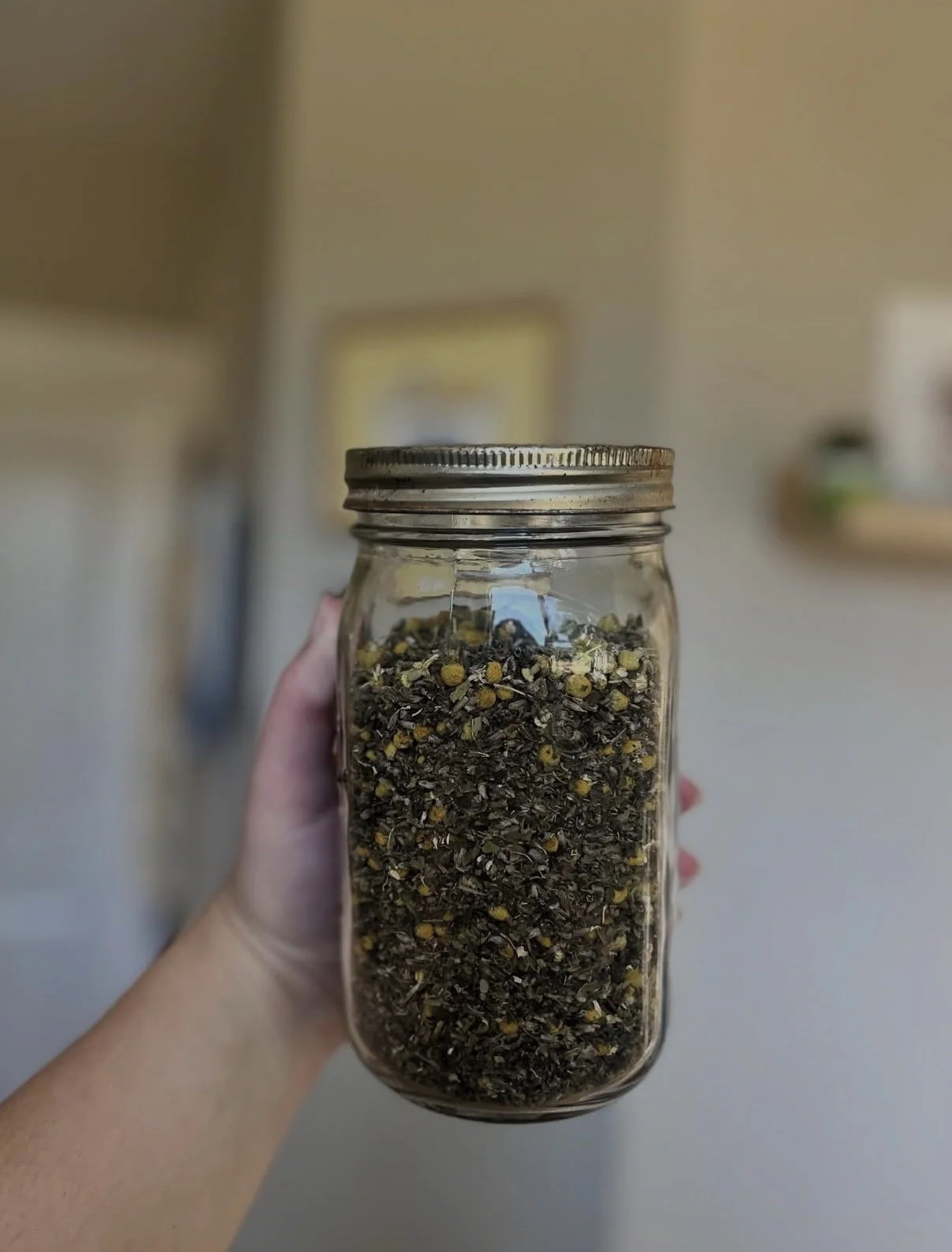 Hand holding a glass jar filled with dried herbs and flowers in a home kitchen.