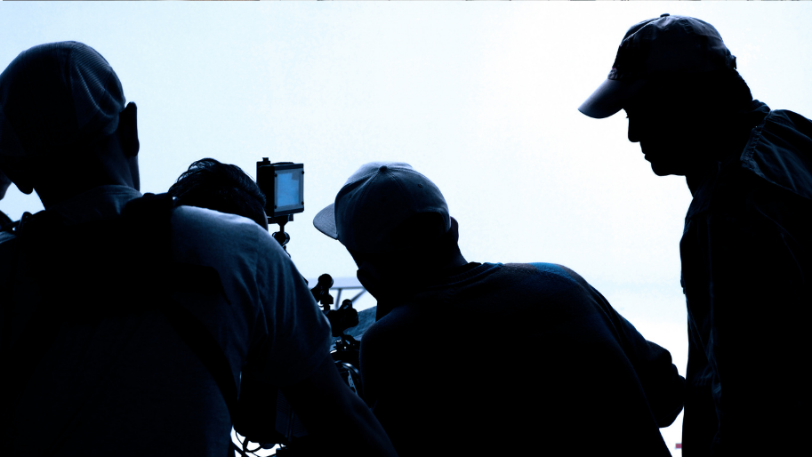 Silhouettes of four people gathered around a camera, likely filming or photographing, against a bright sky.
