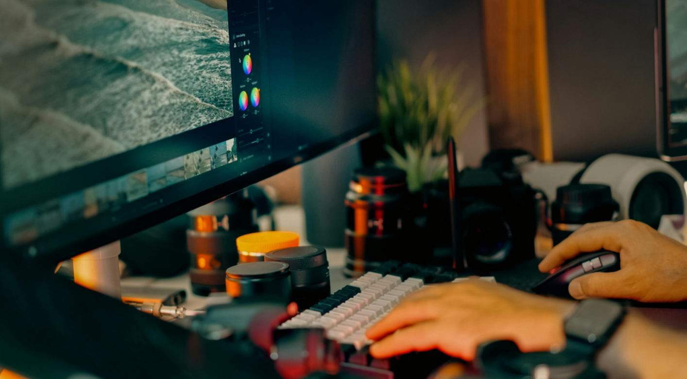 Close-up of a person editing photos on a large computer monitor, with a keyboard, mouse, and various camera lenses and equipment on the desk.