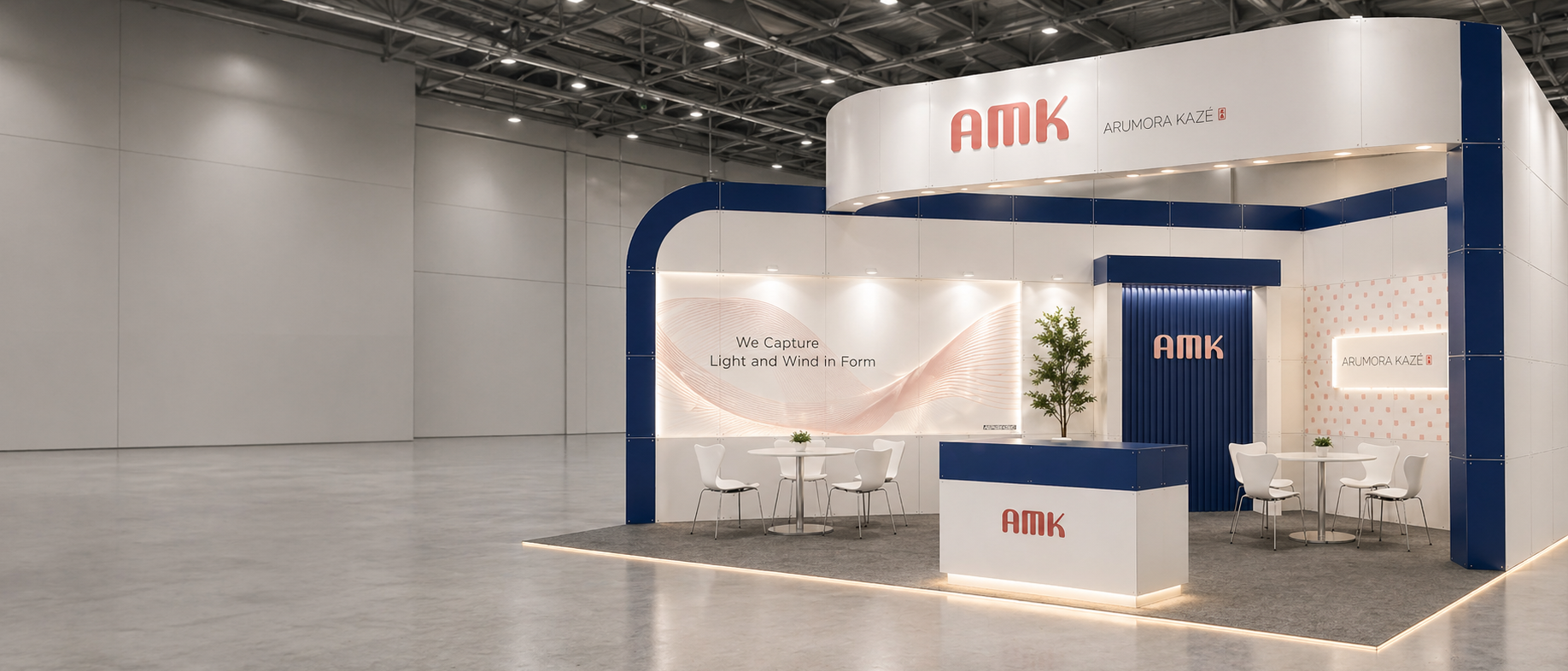 전시 부스 내부 모습, 하단에는 'AMK' 로고가 있고, 소파와 테이블, 화분이 놓여 있으며, 벽에는 'We Capture Light and Wind in Form' 문구가 있다.