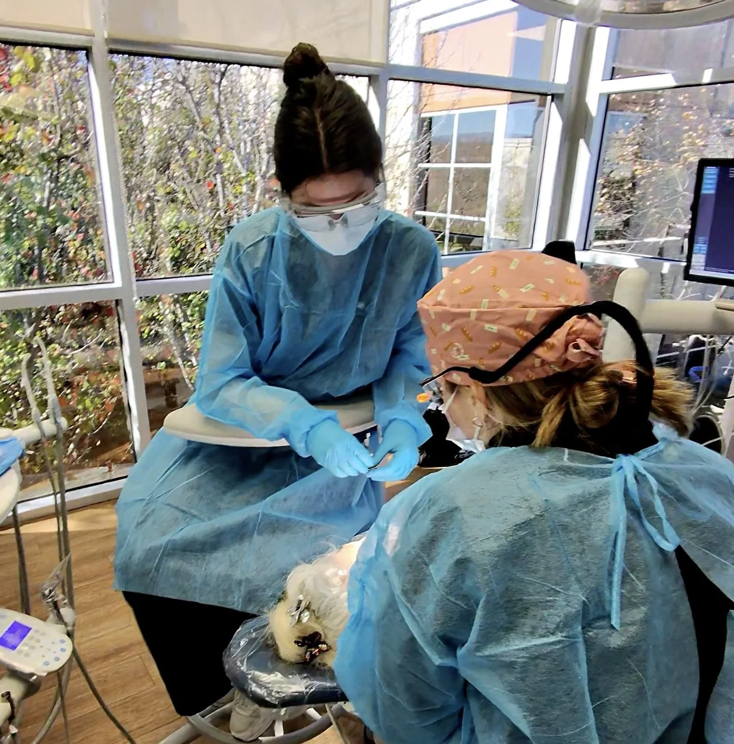 Two medical professionals, one wearing protective goggles and a pink surgical cap, are performing a procedure on a small animal, possibly a dog, in a clinical setting with large windows showing outside foliage.