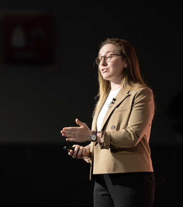 A woman with glasses and long brown hair wearing a beige blazer, white top, and black pants giving a presentation, holding a remote in her right hand and gesturing with her left hand, with a dark background.
