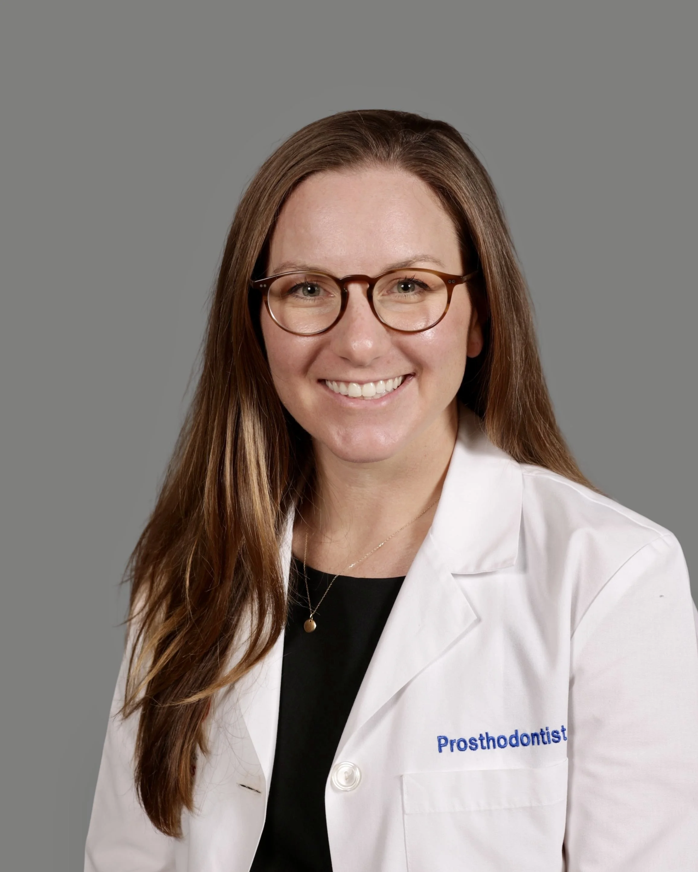 Close-up of a smiling woman with long brown hair, glasses, wearing a white coat with the word 'Prosthodontist' embroidered on it. Background is plain gray.