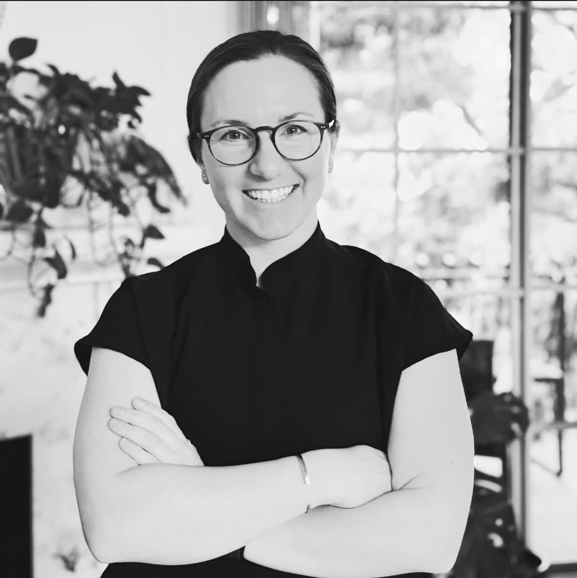 Black and white photo of a smiling woman with glasses, arms crossed, in an indoor space with large windows and plants in the background.