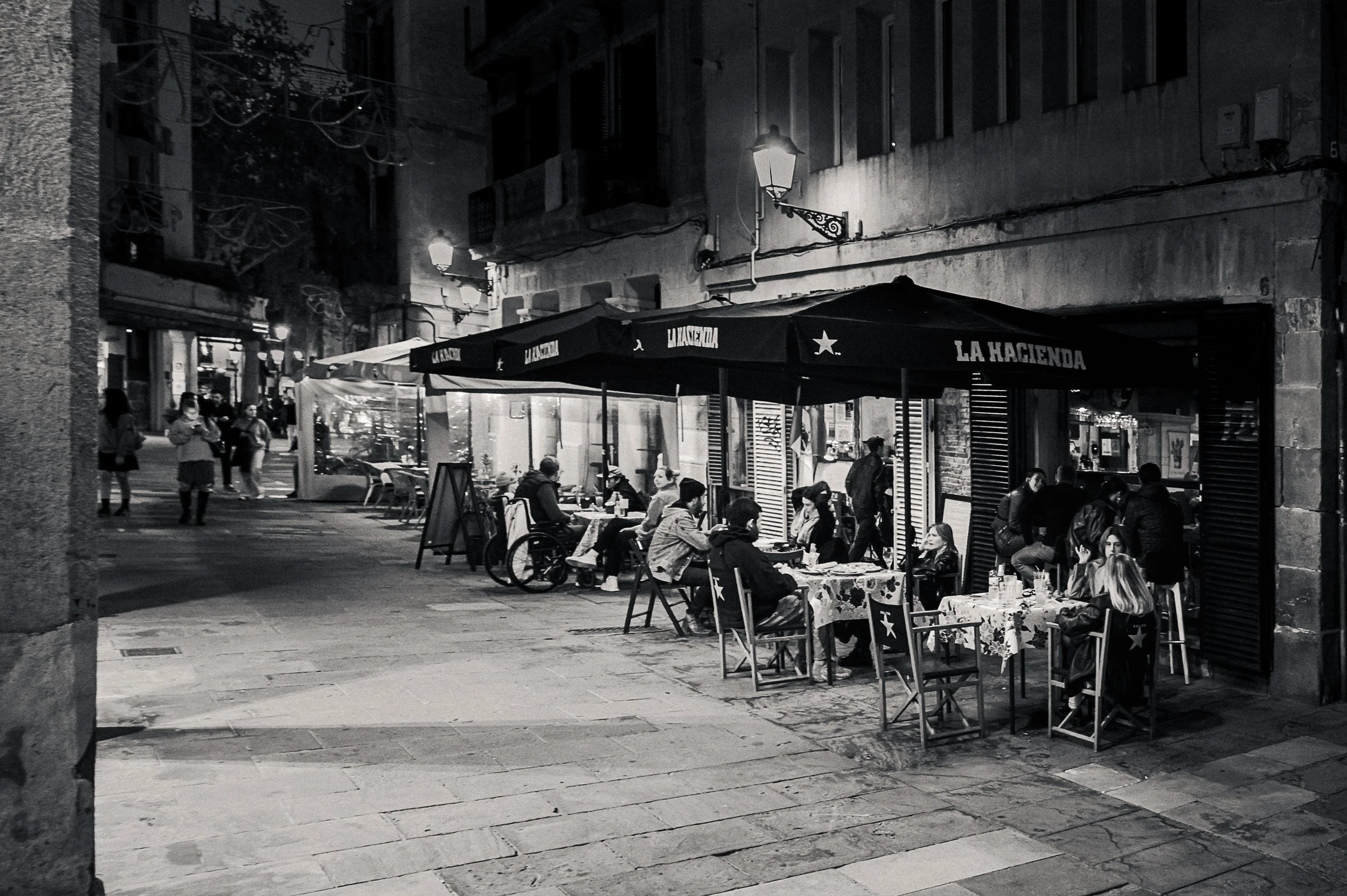 Guests seated outside a Barcelona street café under warm lamps at night.