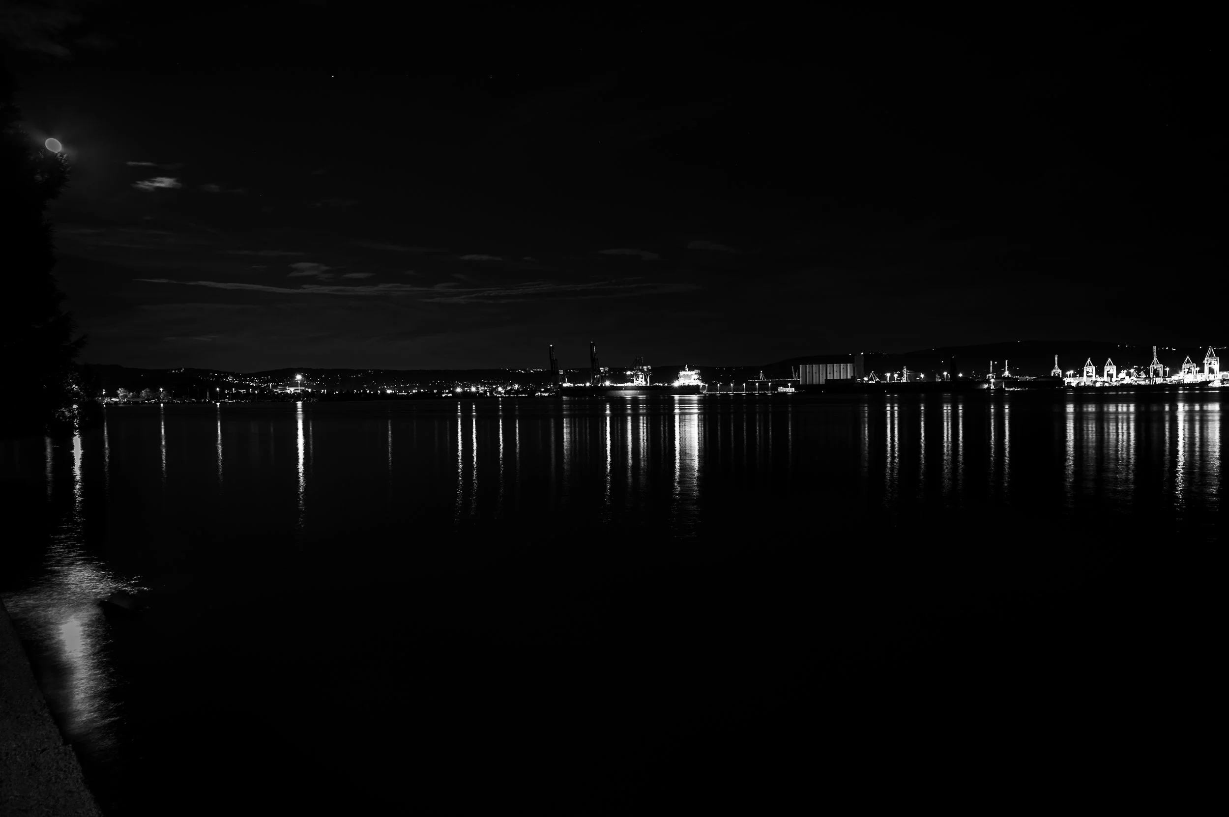 Moon above an industrial harbour at night, with cranes and long reflections across calm water.
