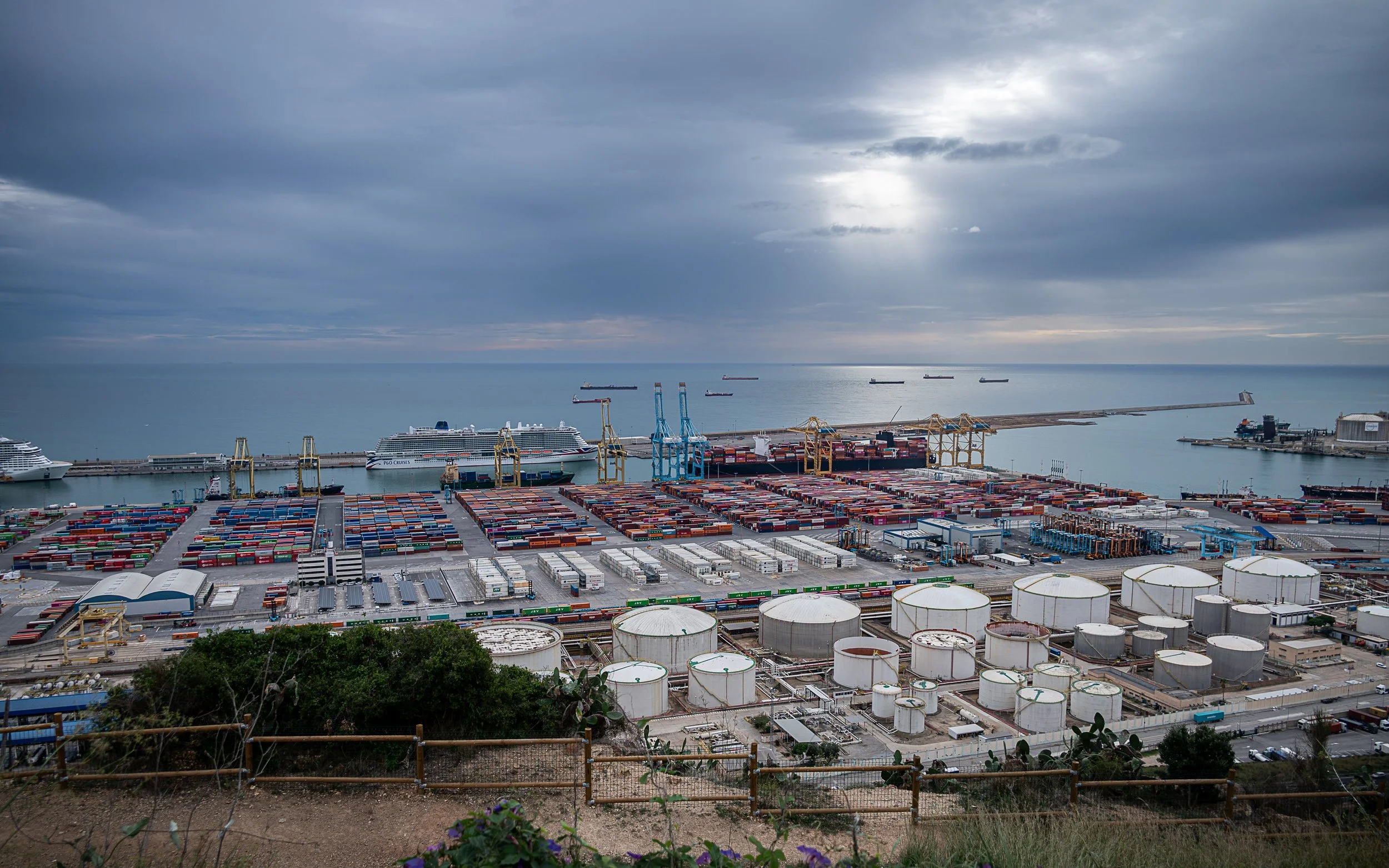 Barcelona port with containers, storage tanks and ships beneath a dramatic cloudy sky.