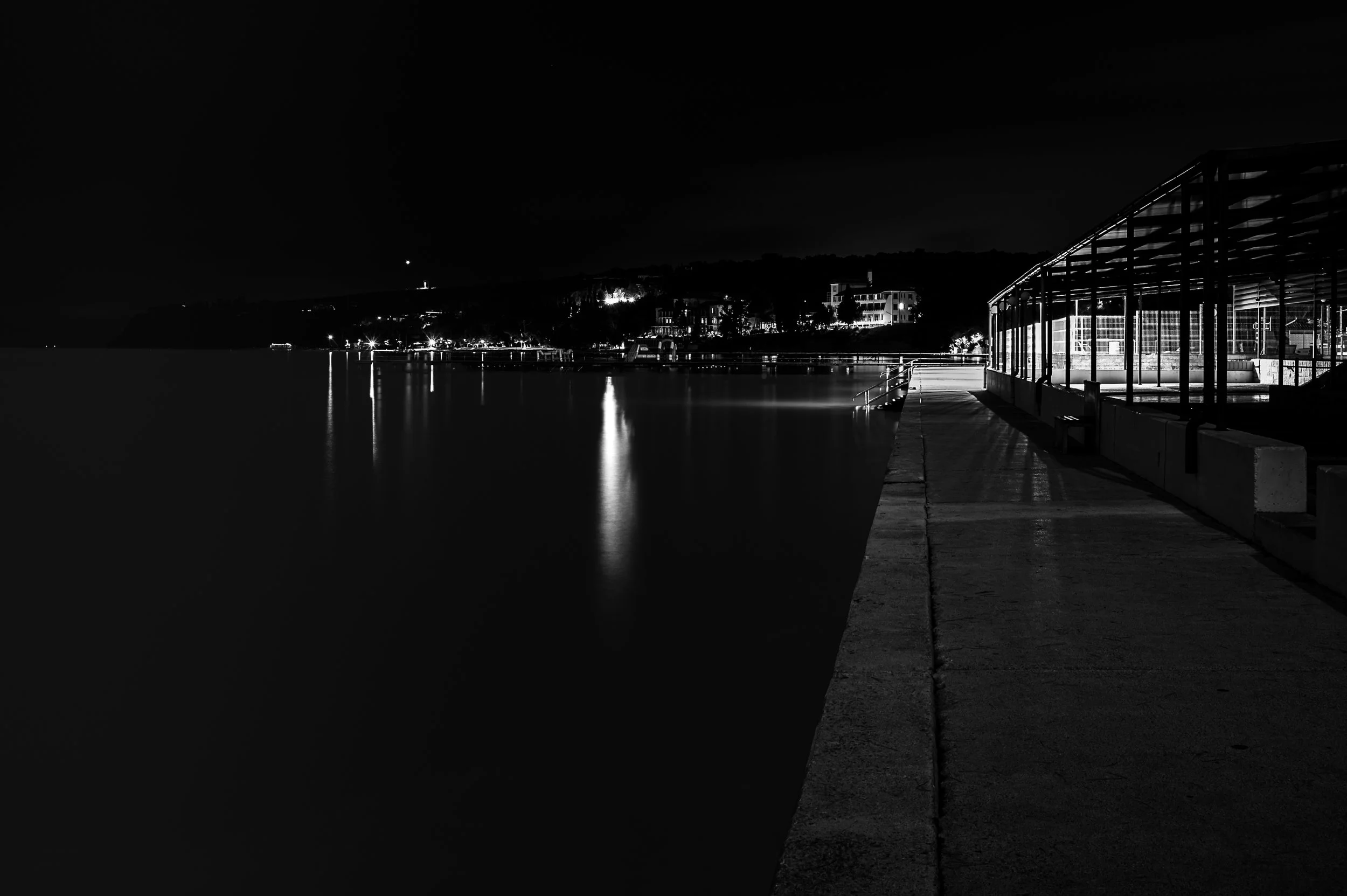 Concrete waterfront promenade leading toward distant lights reflected on calm water at night.