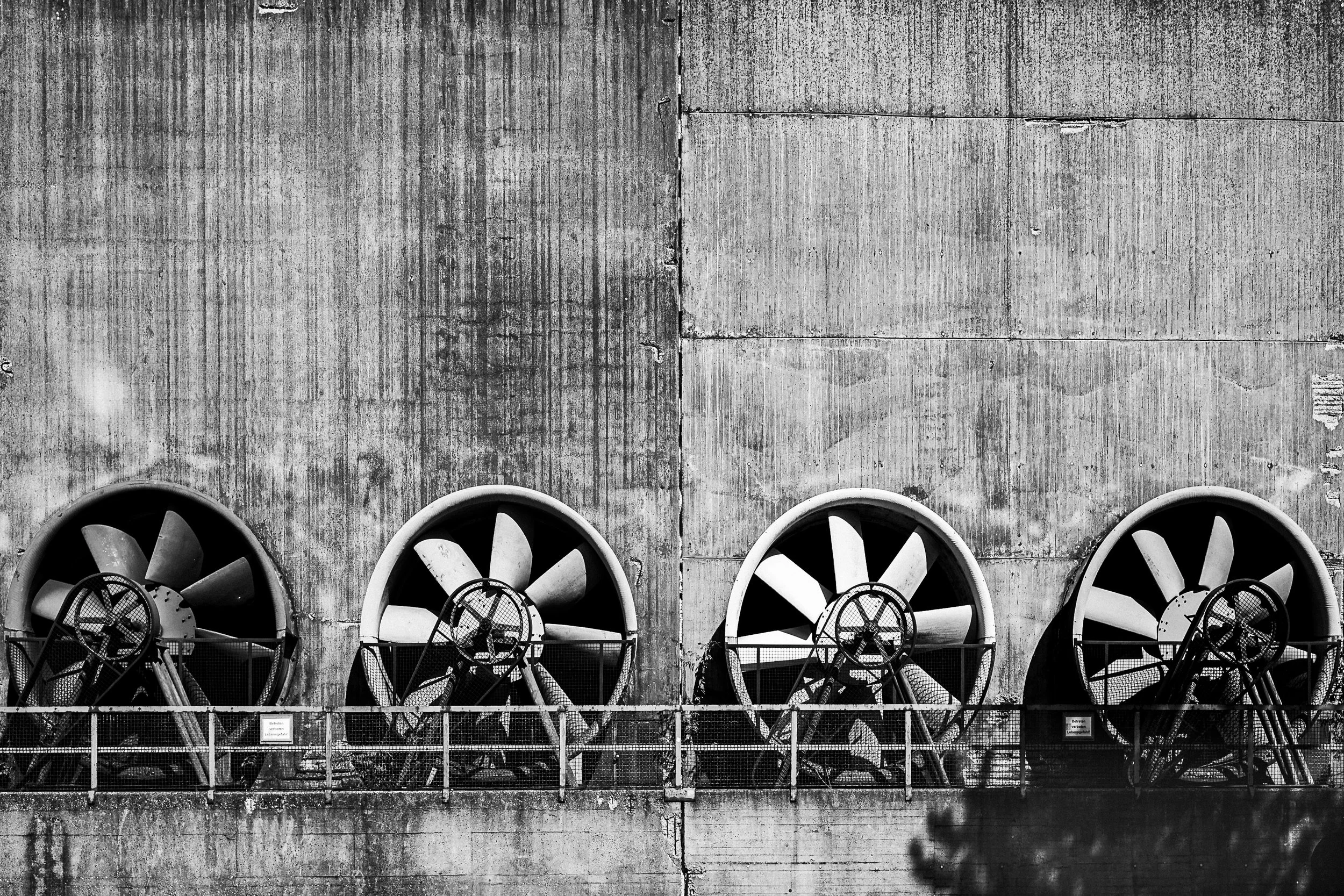 Four large ventilation fans repeat across a weathered concrete industrial wall.