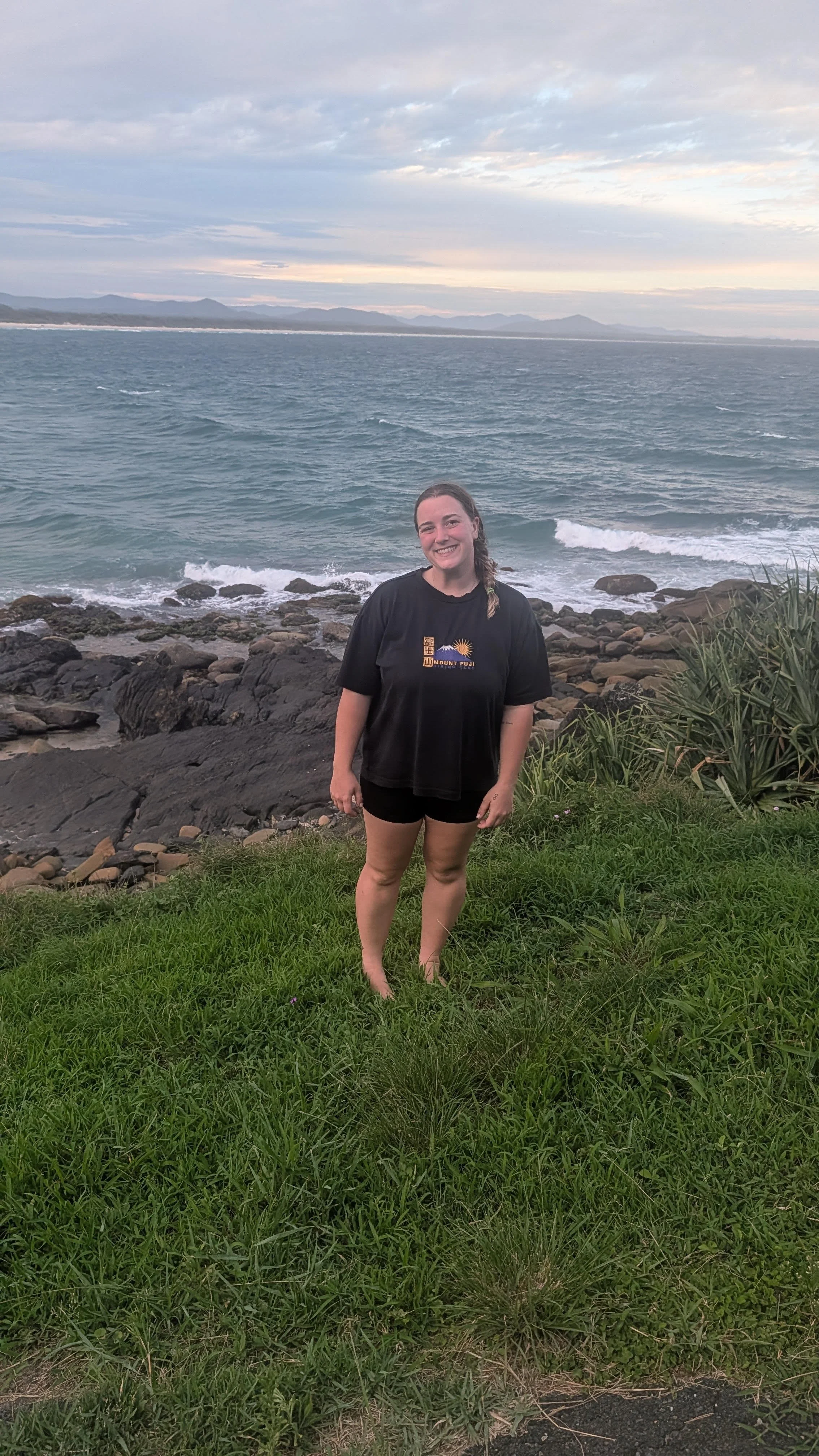A woman smiling at the camera while standing on grass near a rocky shoreline with an ocean and mountains in the background during cloudy weather.