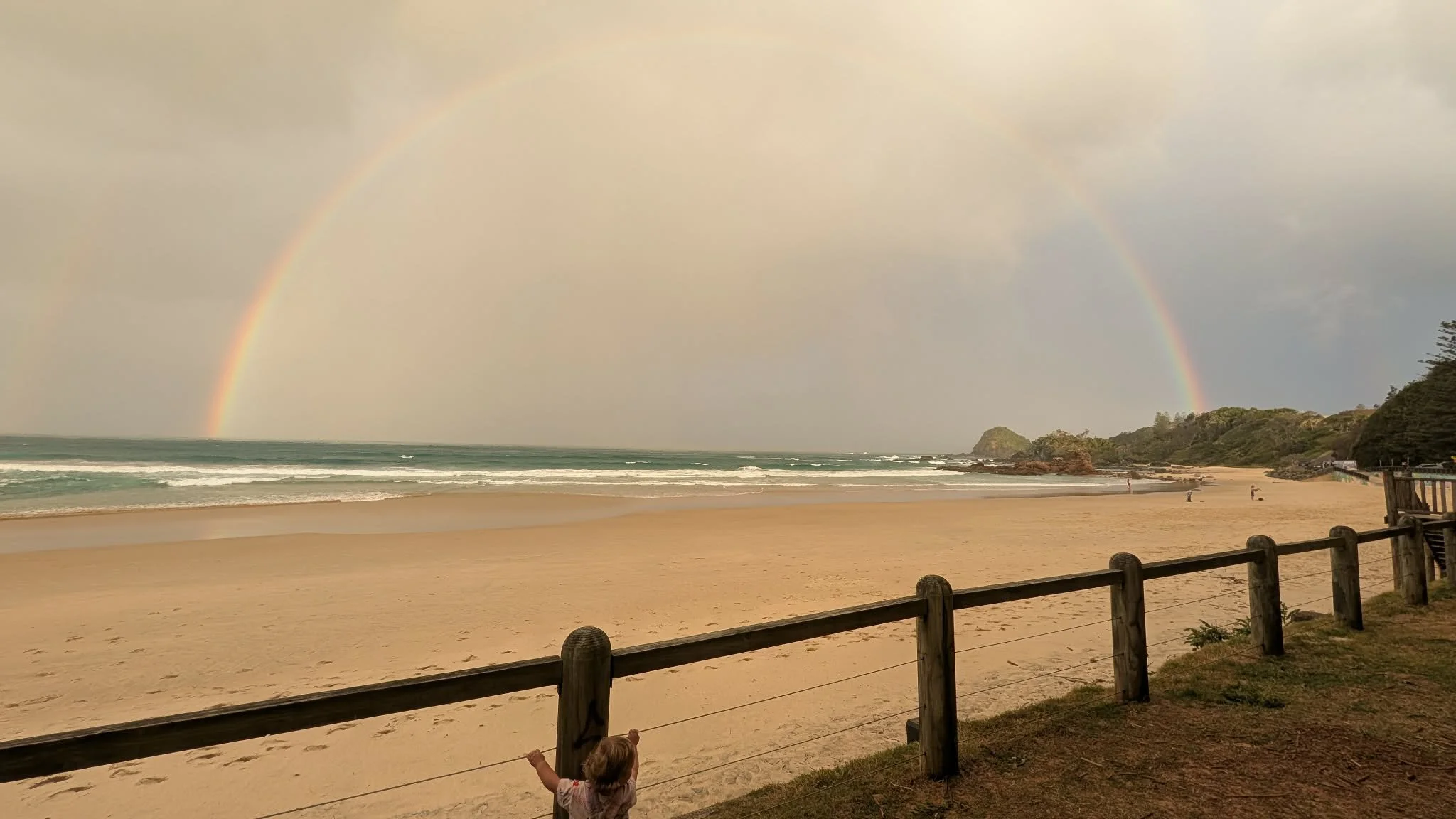 A child standing near a wooden fence on a sandy beach, with a rainbow in the cloudy sky over the ocean and distant hills.