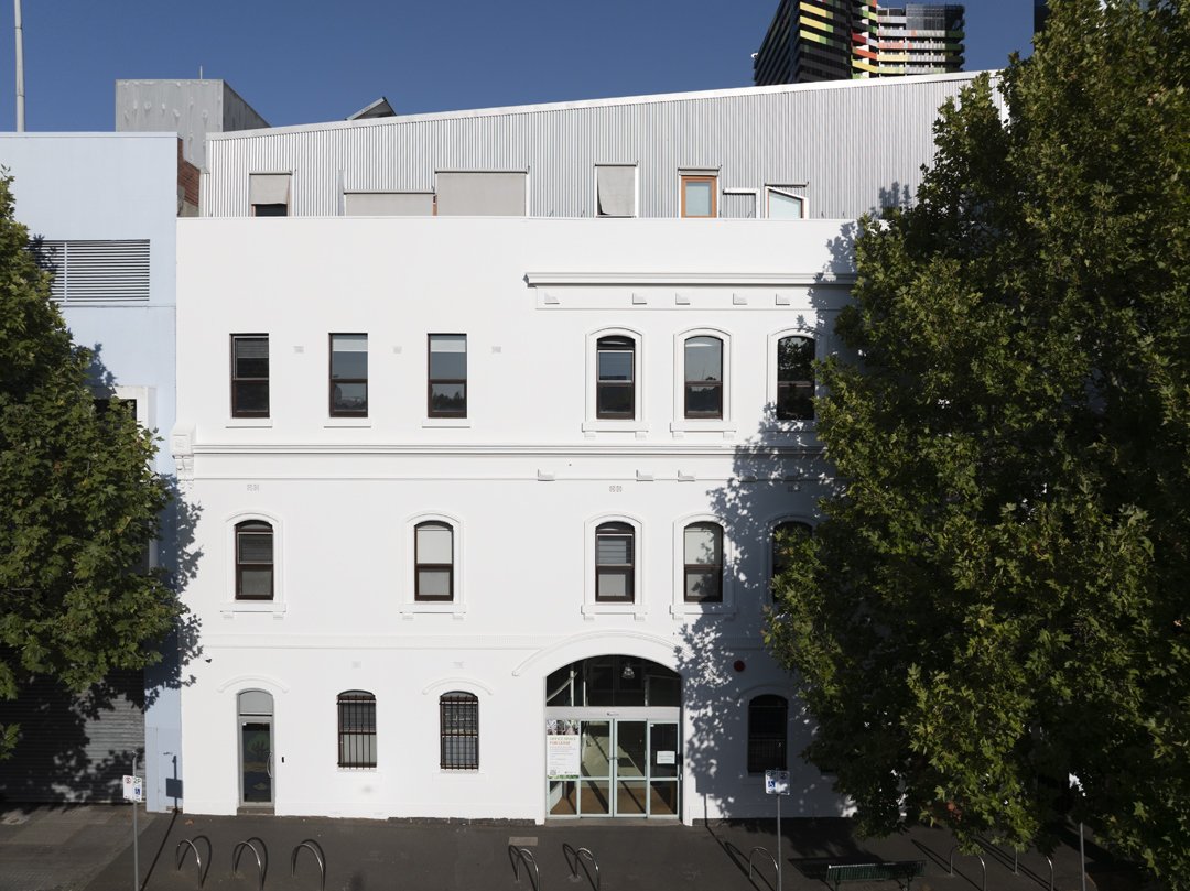 A white multi-story building with arched windows on the ground floor, rectangular windows on upper floors, and some trees in front. The building has a modern and historic architectural style.