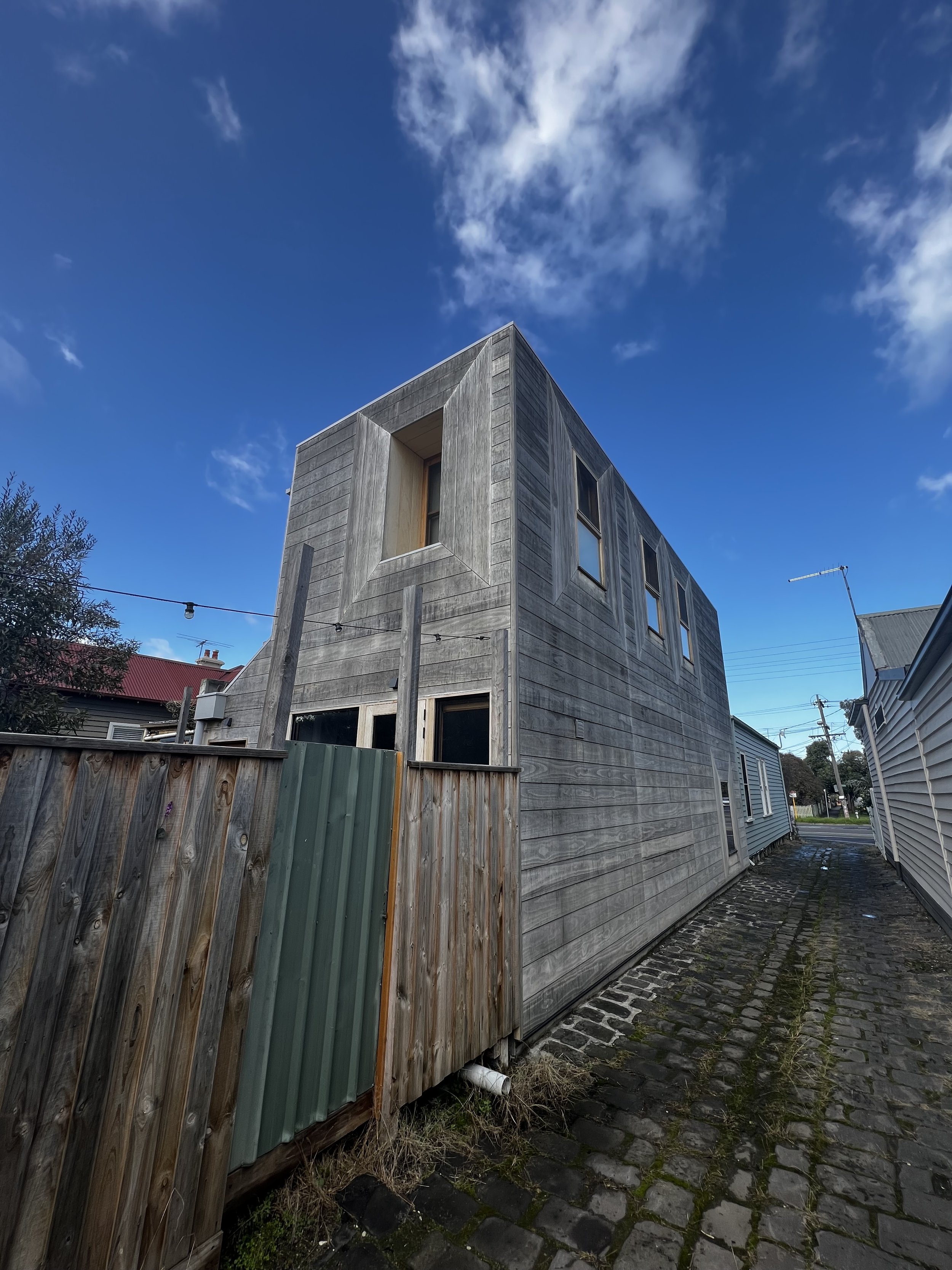 A wooden modern house with multiple rectangular windows under a partly cloudy blue sky, situated along a cobblestone alley with a weathered wooden fence.