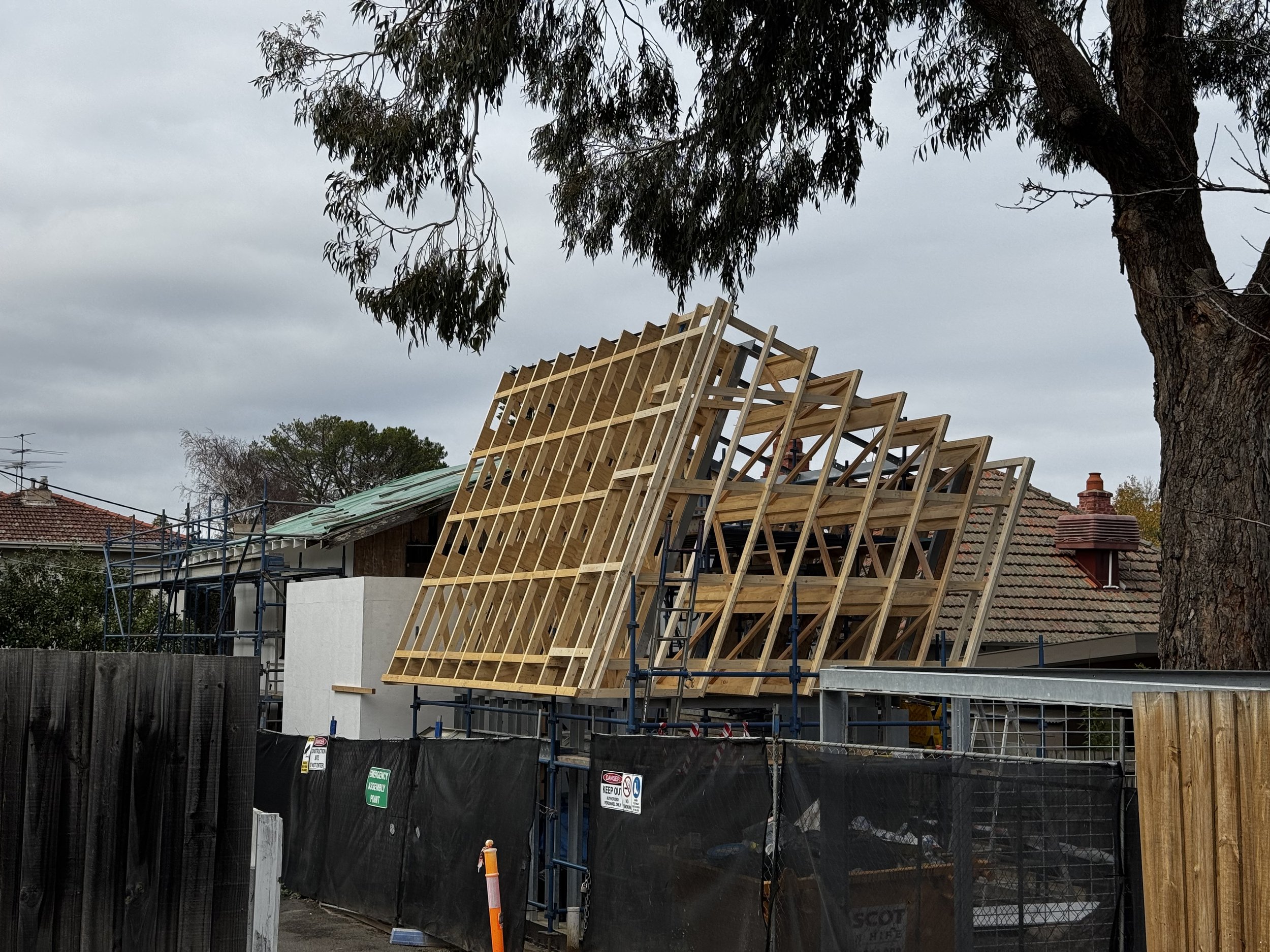 Construction site with wooden framing for a roof and house, scaffolding and black safety barriers, under a cloudy sky, with trees and neighboring houses in the background.