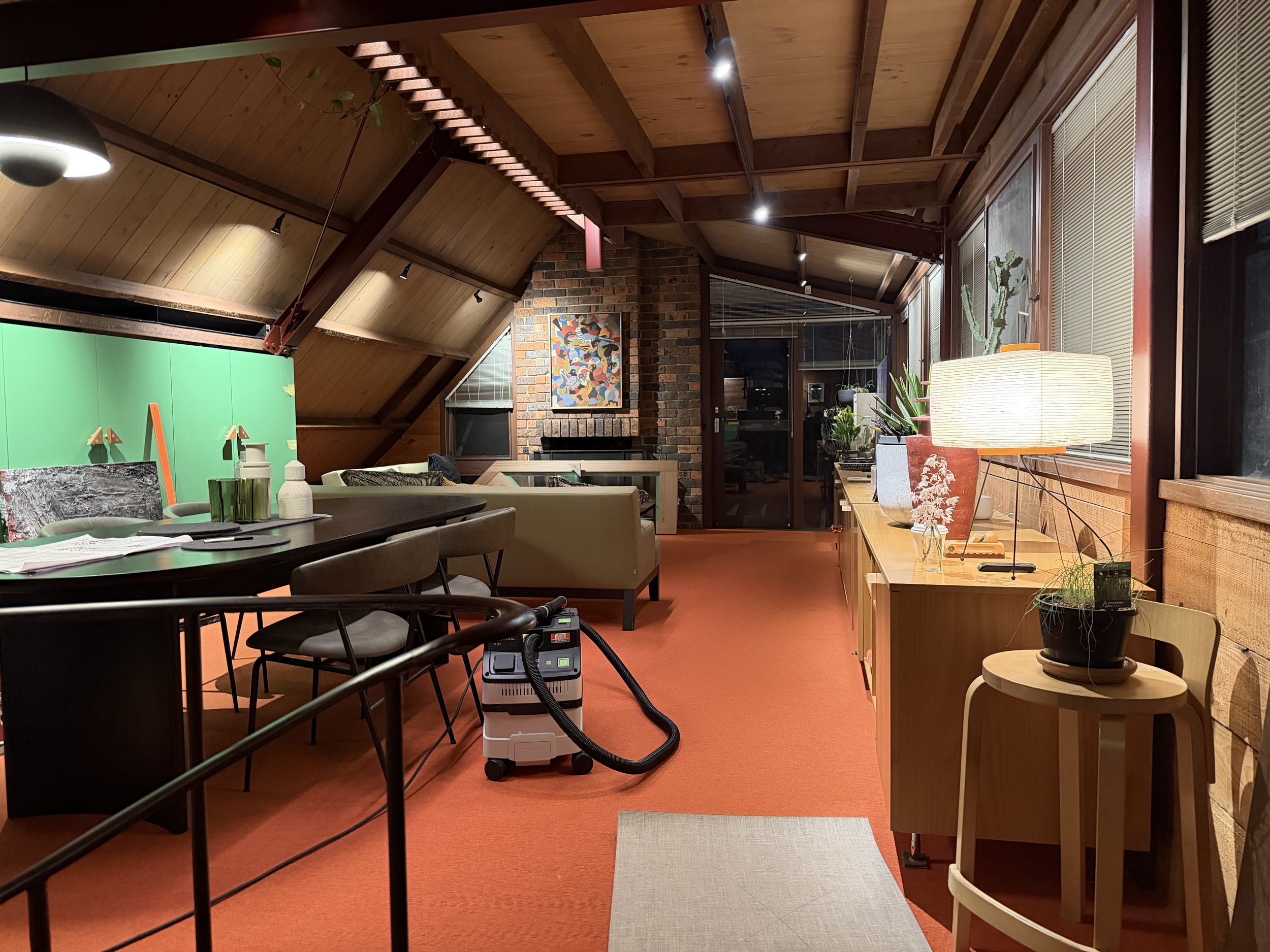 A cozy living space with a wood-paneled ceiling and brick wall, illuminated by warm lighting. Contains a green accent wall, a black table with chairs, a beige sofa, multiple houseplants on a wooden sideboard, and a massage table. A vacuum cleaner is on the red carpeted floor, and artwork decorates the brick wall.