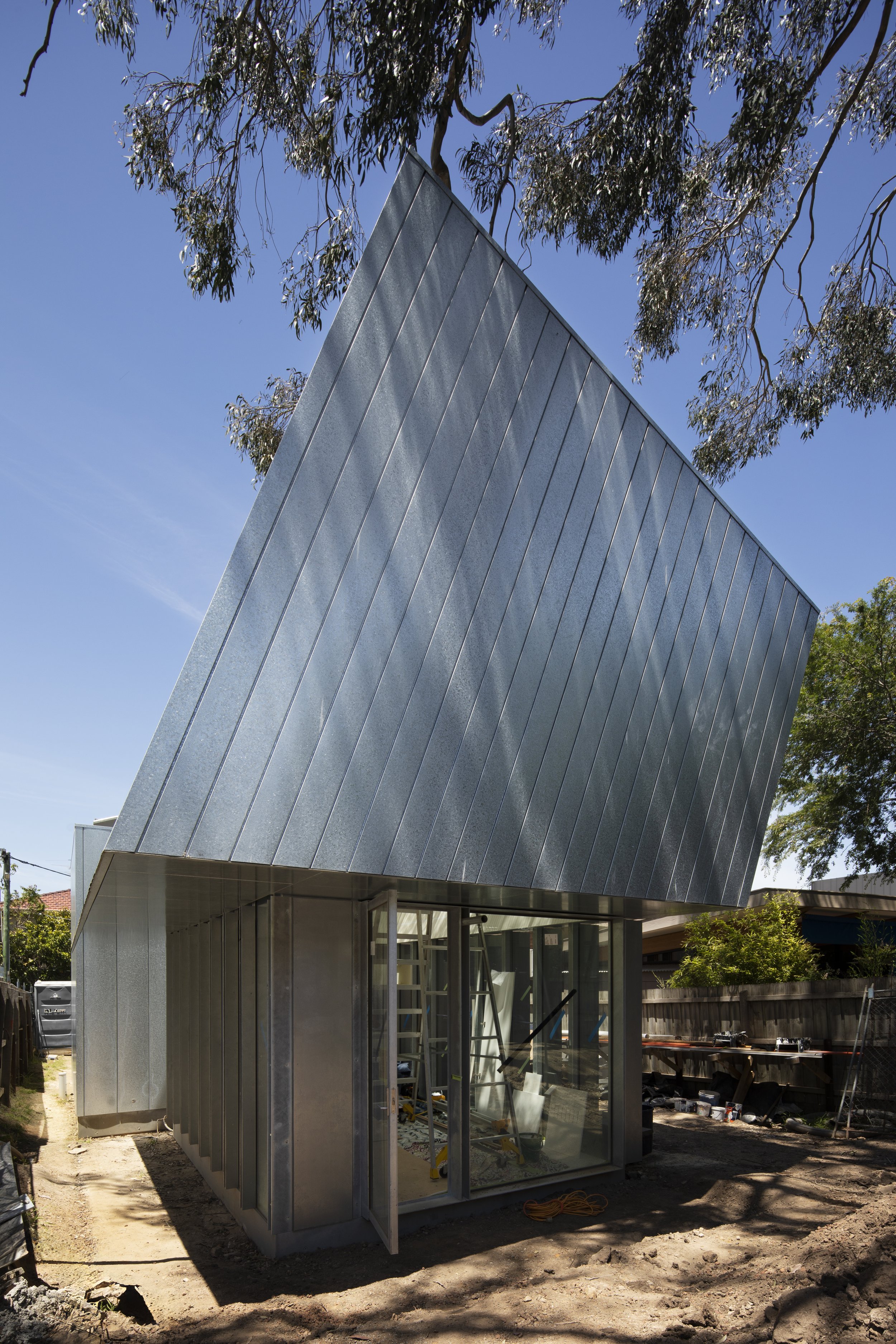 Modern small building under construction with metallic exterior and large glass doors, surrounded by trees and construction tools.