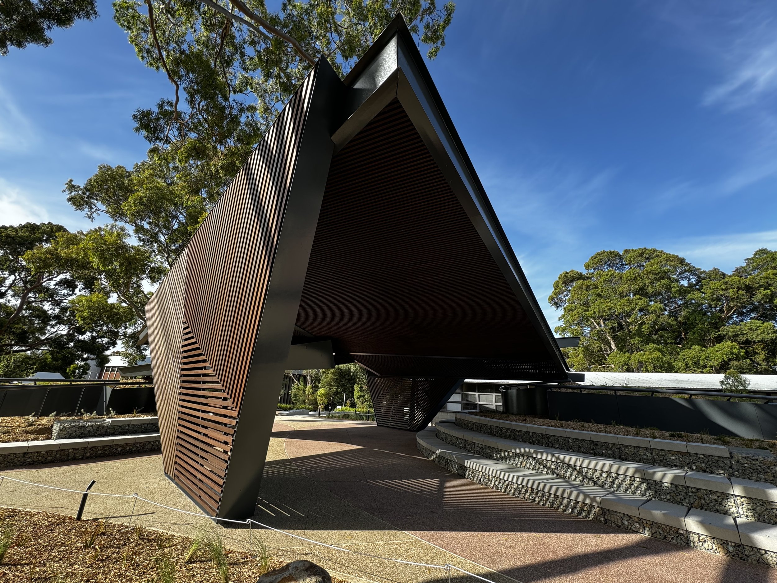 Modern building with angular, overhanging roof and wooden slat facade, surrounded by trees and a landscaped area
