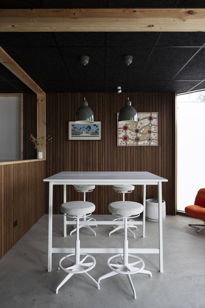 Modern dining area with a high white table, three white bar stools, dark wood-paneled walls, two hanging pendant lights, abstract wall art, and a red chair near a large window.