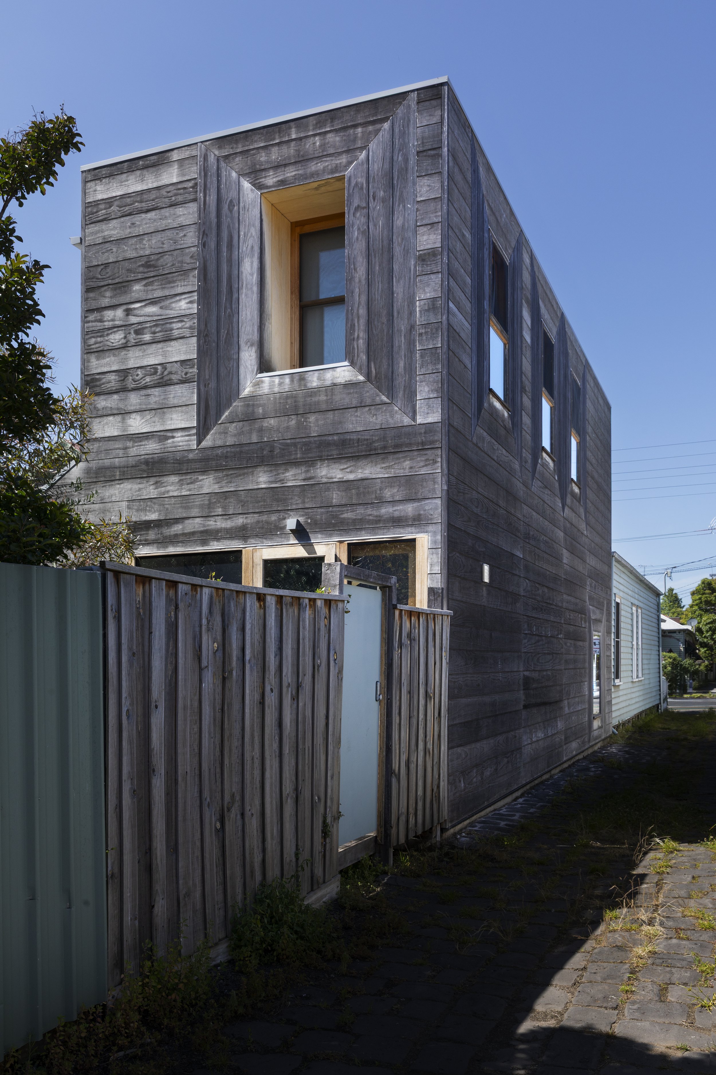 A modern, two-story house with weathered wooden exterior siding and multiple windows, enclosed by a wooden fence with a frosted glass gate, situated on a narrow cobblestone path under a clear blue sky.