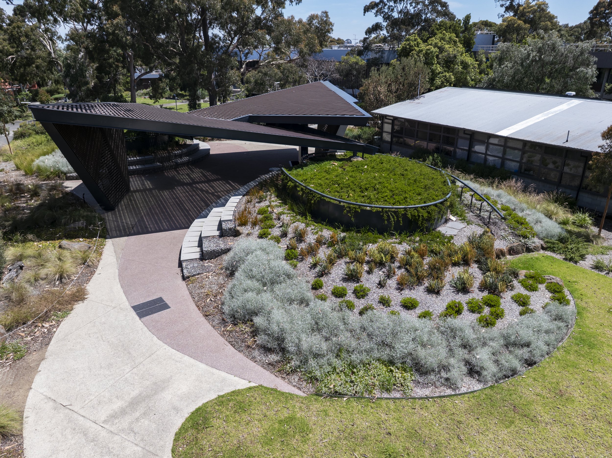 A modern building with a uniquely angled roof and an outdoor seating area, surrounded by landscaped garden beds with various plants and a curved pathway in a park-like setting.