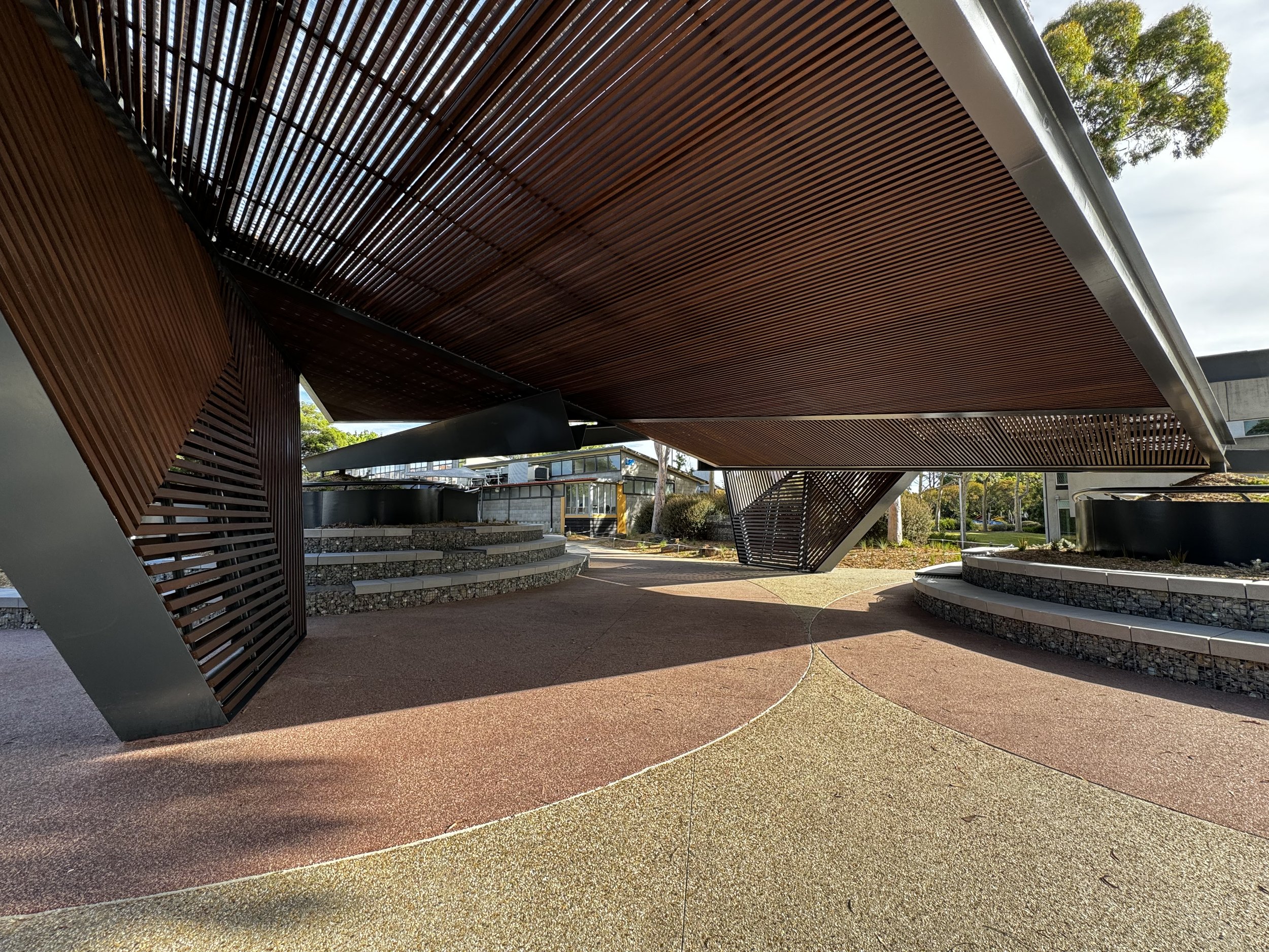 Modern architectural structure with large overhanging roof supported by angled beams, featuring decorative wooden slats, surrounded by landscaped pathways and plants.