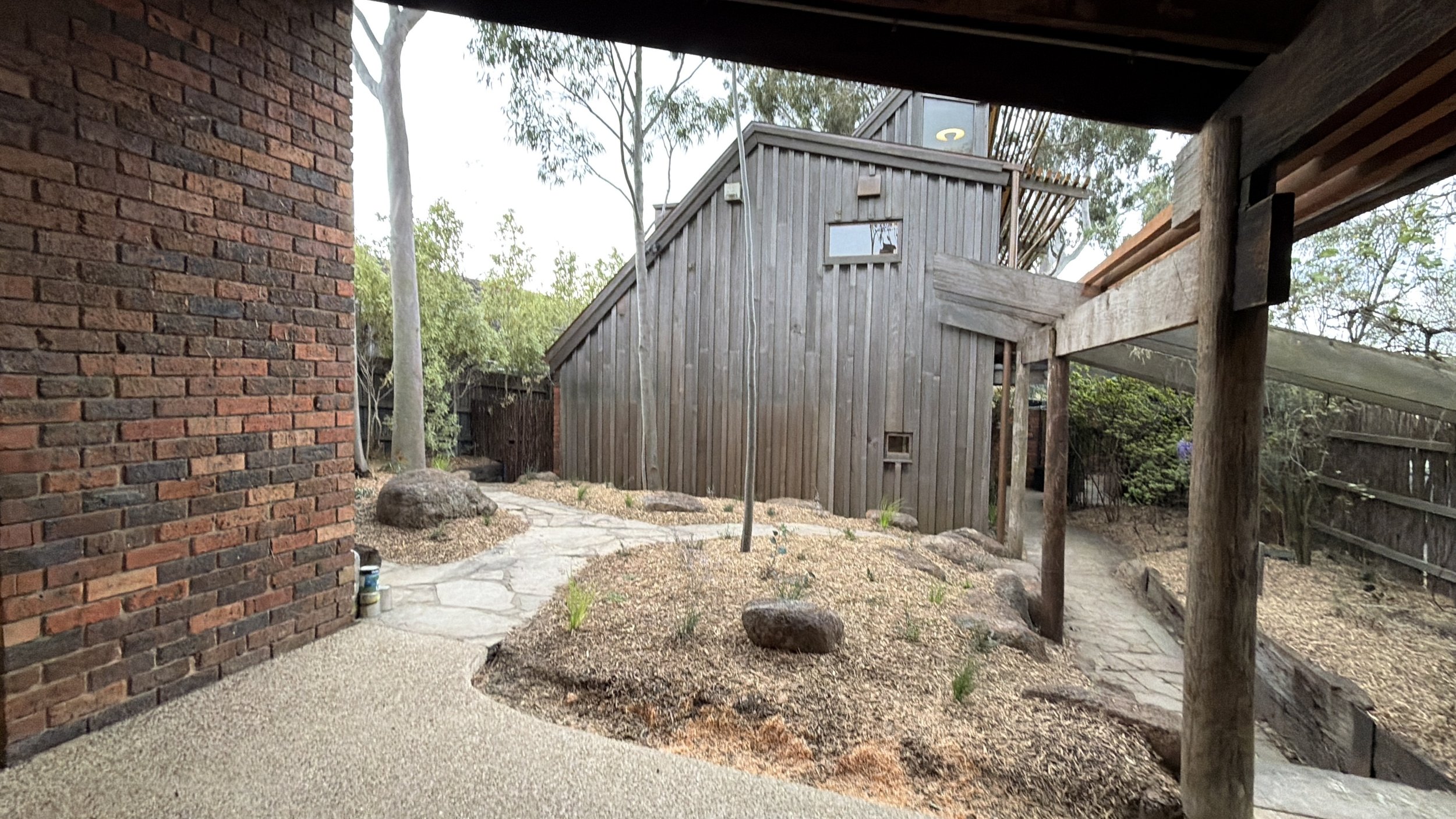 Backyard area featuring a brick wall on the left, a stone pathway, an area with small plants and rocks, and a wooden house with a sloped roof in the background. Foliage and trees are also visible.
