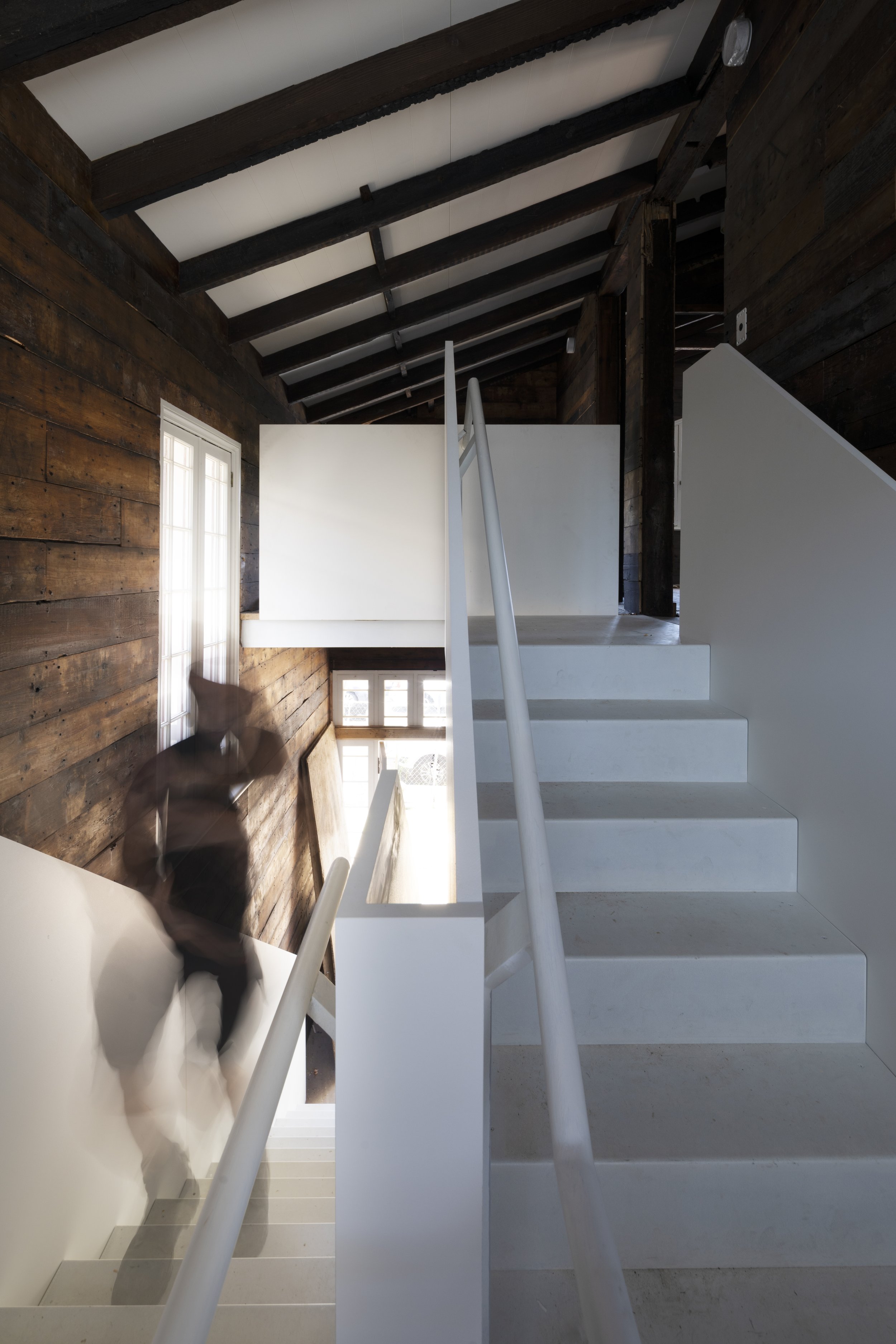 Interior view of a modern stairway with white stairs and metal railings, wooden wall panels, large windows, and a person in motion blurry descending the stairs.