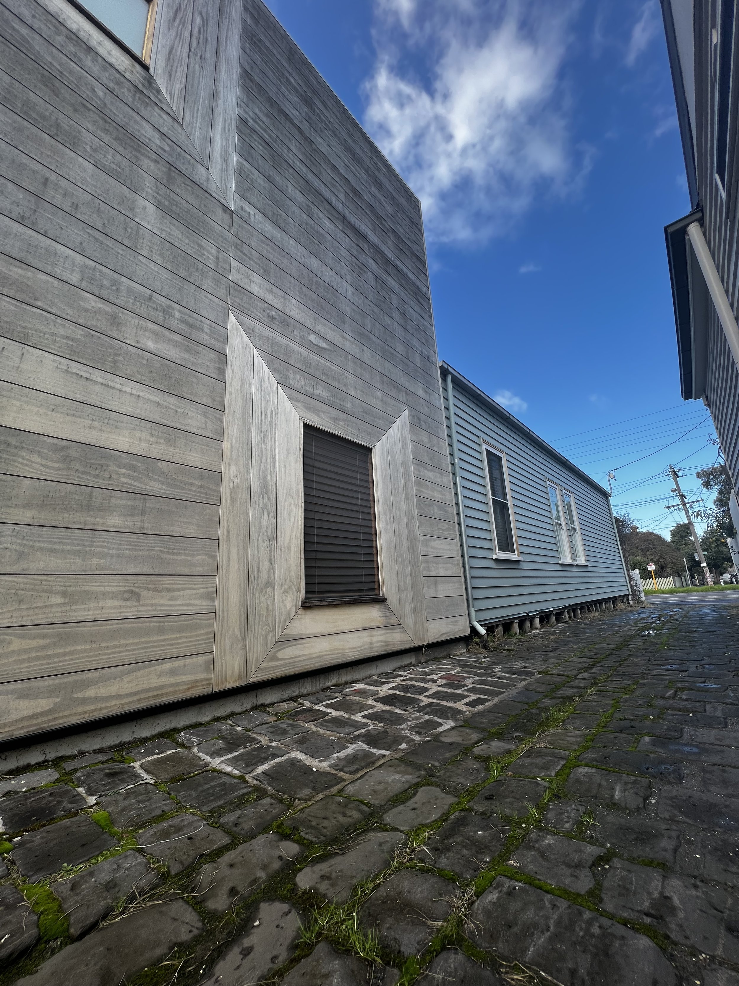 Low-angle view of two houses with blue sky and clouds, featuring weathered wood and vinyl siding, on a brick sidewalk with moss and grass growing between the bricks.